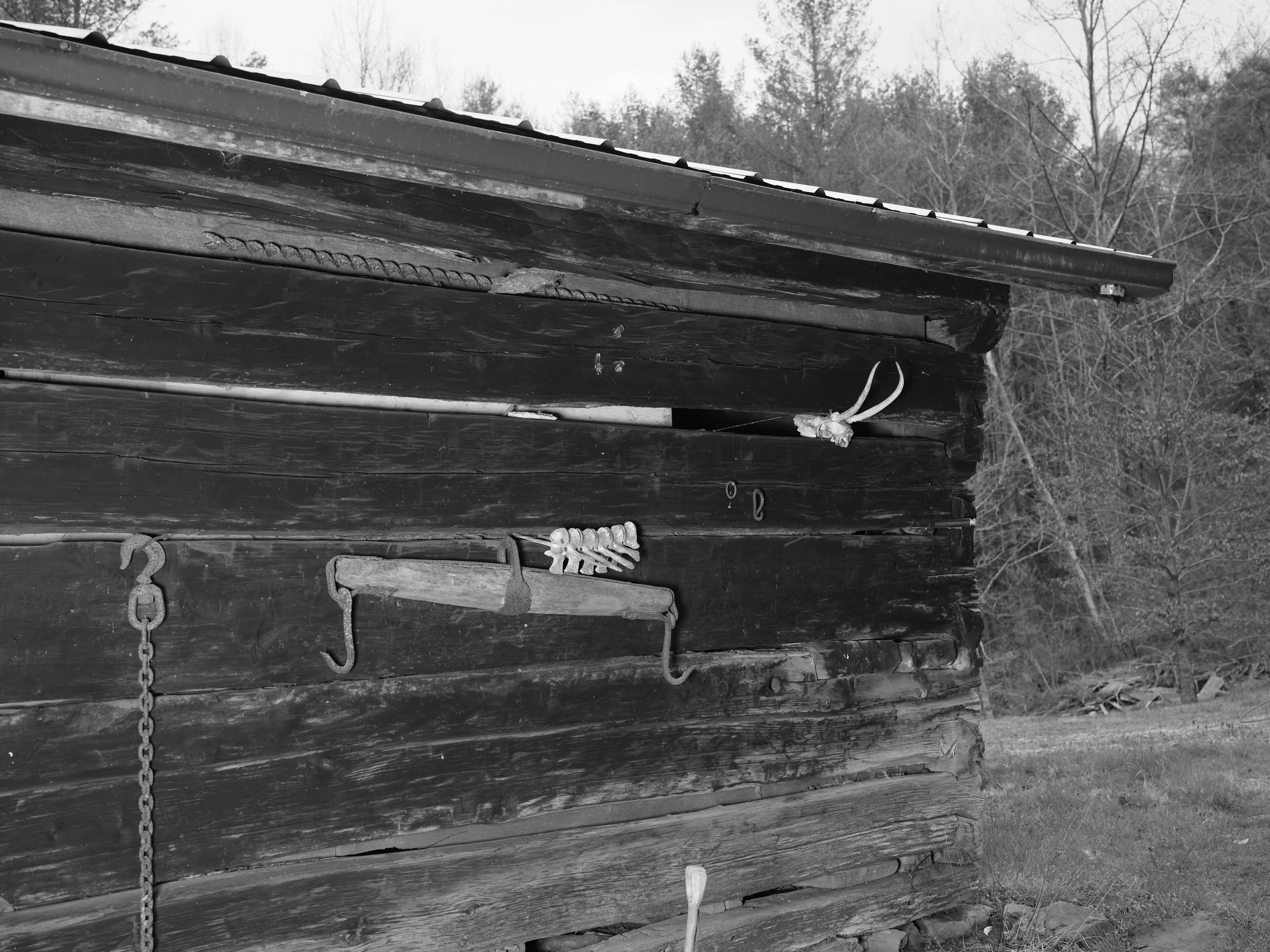 Black-and-white photo of an old wooden building with metal hooks and a chain hanging on the side, and trees in the background.