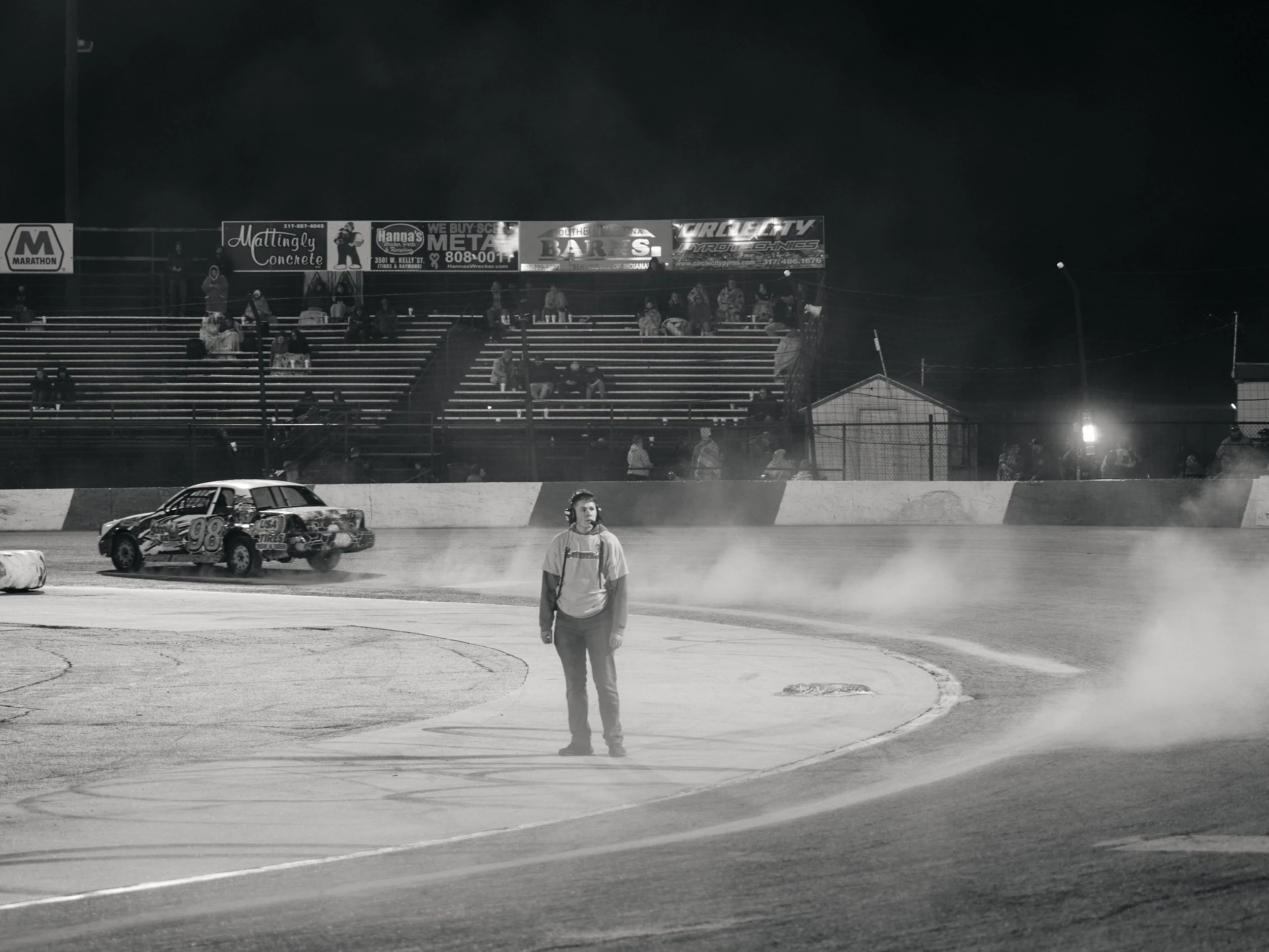 A nighttime scene at a race track shows a person standing in the middle of the track wearing a headset. A race car with the number 99 is on the track, kicking up dust or smoke. Spectators are seated in the bleachers in the background, and advertisements are visible above the bleachers.