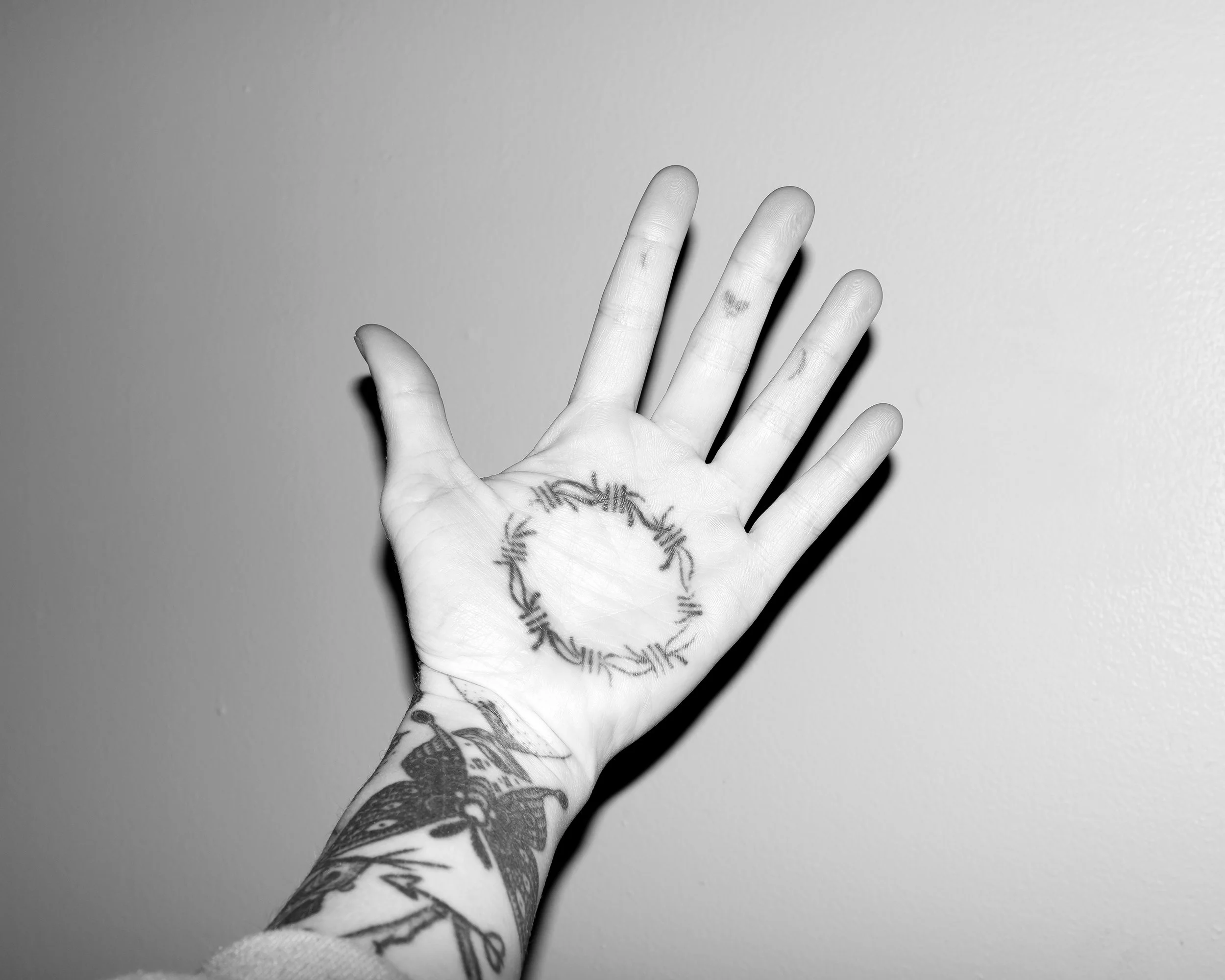 Black and white photo of a human hand with tattoos, holding up against a plain wall background. The hand shows a circular tattoo on the palm and various tattoos on the wrist and forearm.