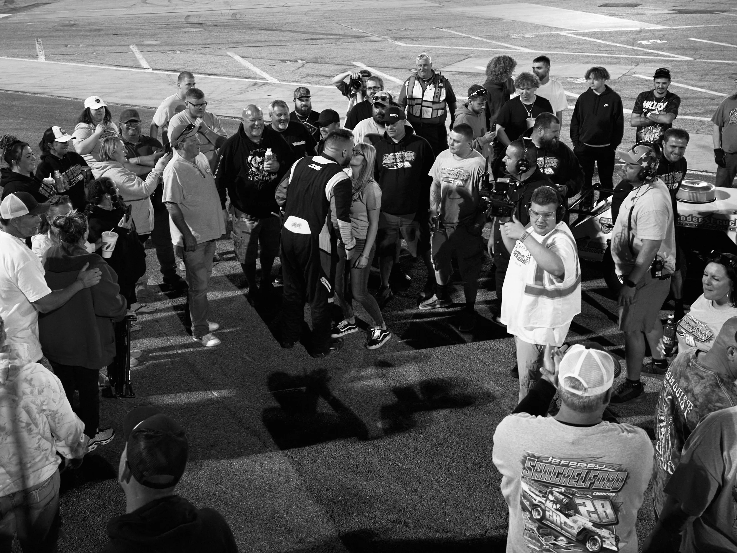 A group of people gathered around a race car on a track at night, with some taking photos and videos, and some wearing racing or casual attire.