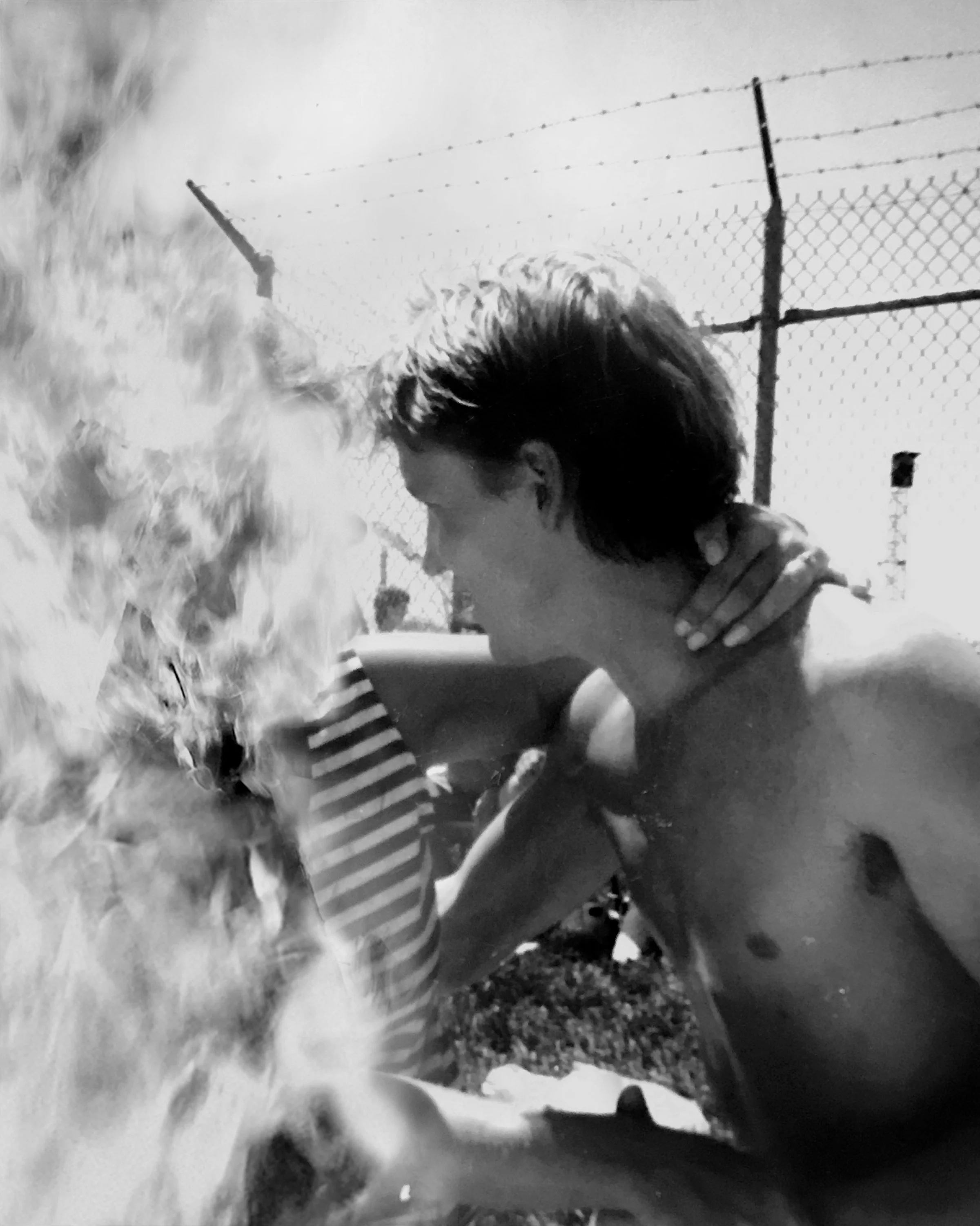 A shirtless man with dark hair and a woman in a striped shirt near a chain-link fence, with a cloud of smoke or dust nearby.