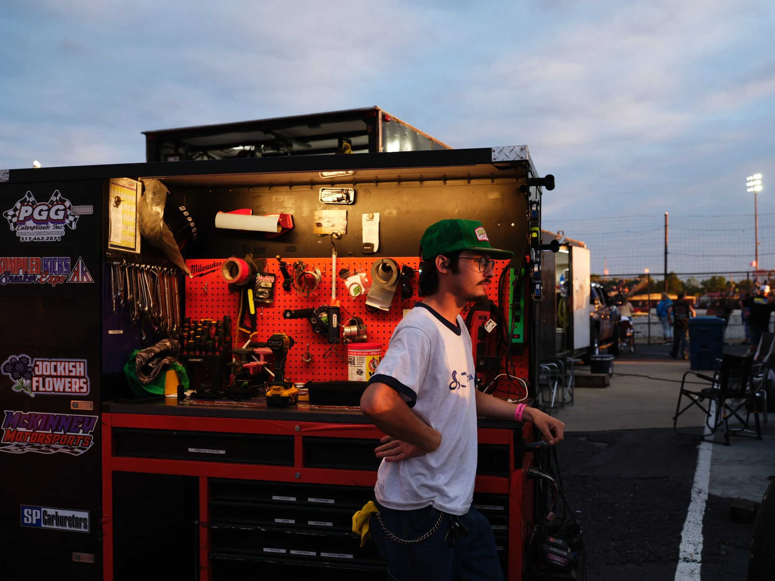 A young man with glasses and a green cap stands in profile beside a red tool chest at a racing event. In the background, there are people, cars, and a fenced race track under a partly cloudy sky during dusk.