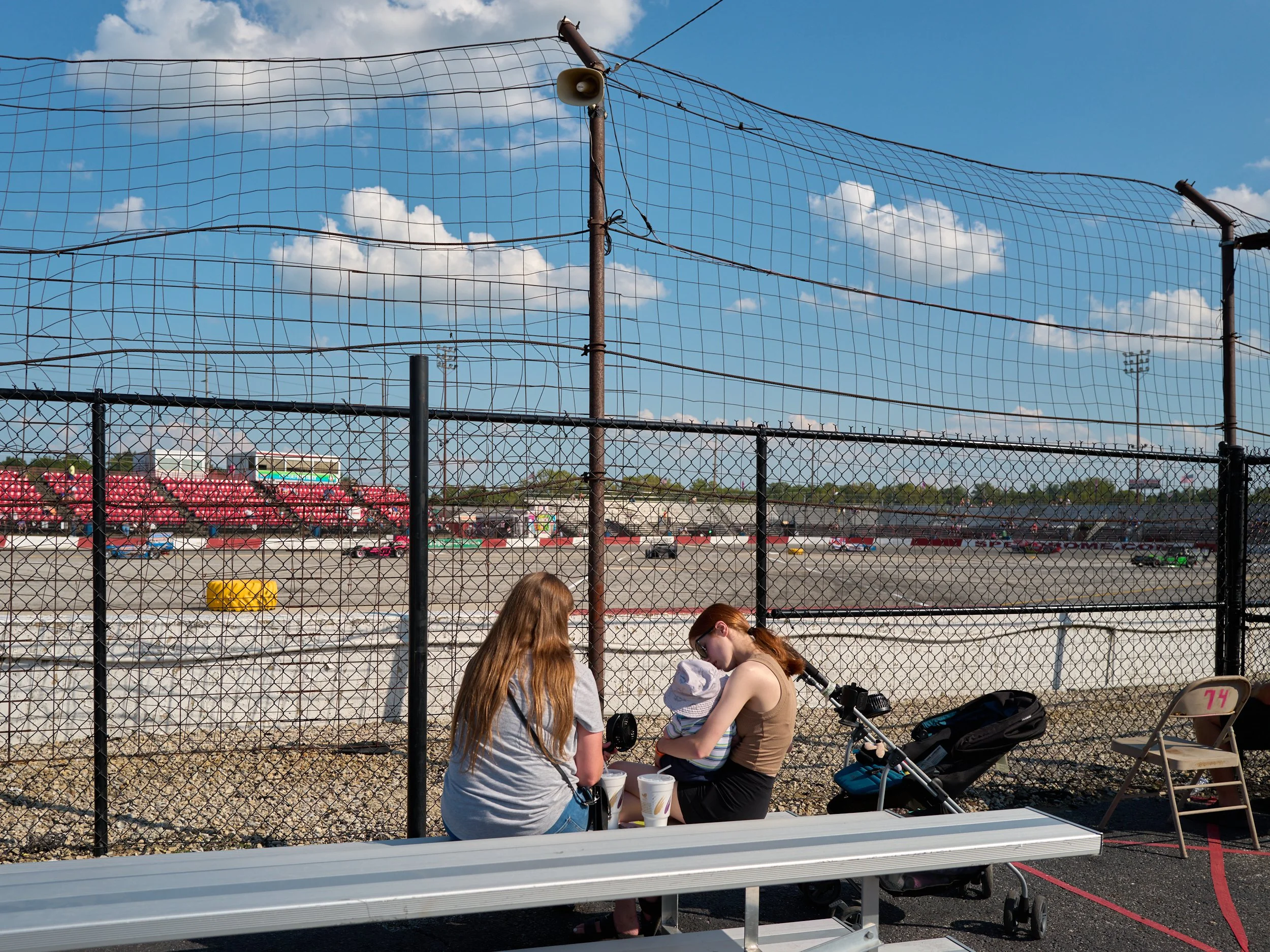 Two women sitting on a white park bench near a chain-link fence at a racetrack, one with long hair and the other holding a baby, with a stroller nearby on a sunny day.
