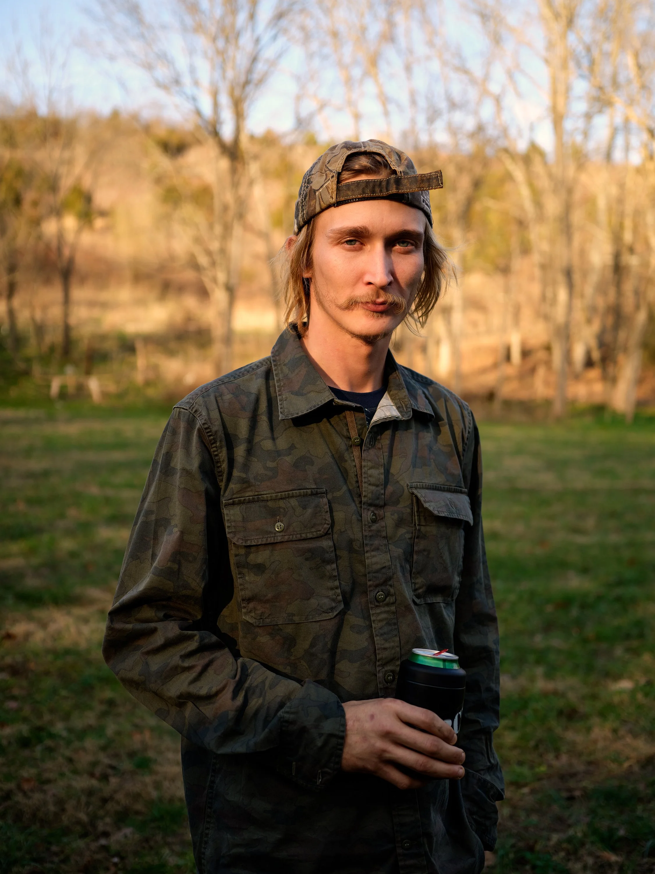 A young man with shoulder-length blonde hair, a mustache, and a goatee, standing outdoors in a wooded area during late afternoon or early evening. He is wearing a camouflage jacket, a camouflage baseball cap worn backwards, and holding a black can koozie with a beverage can inside.