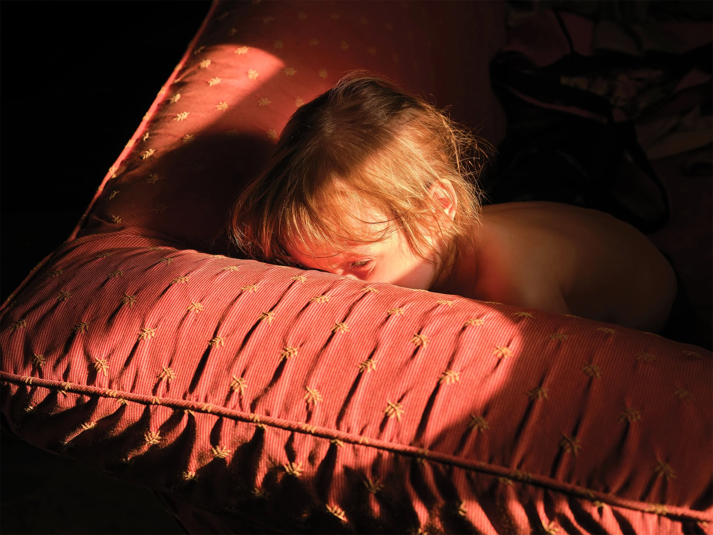 A child lying on a patterned bed in warm lighting, with hair illuminated by sunlight and face partially obscured.