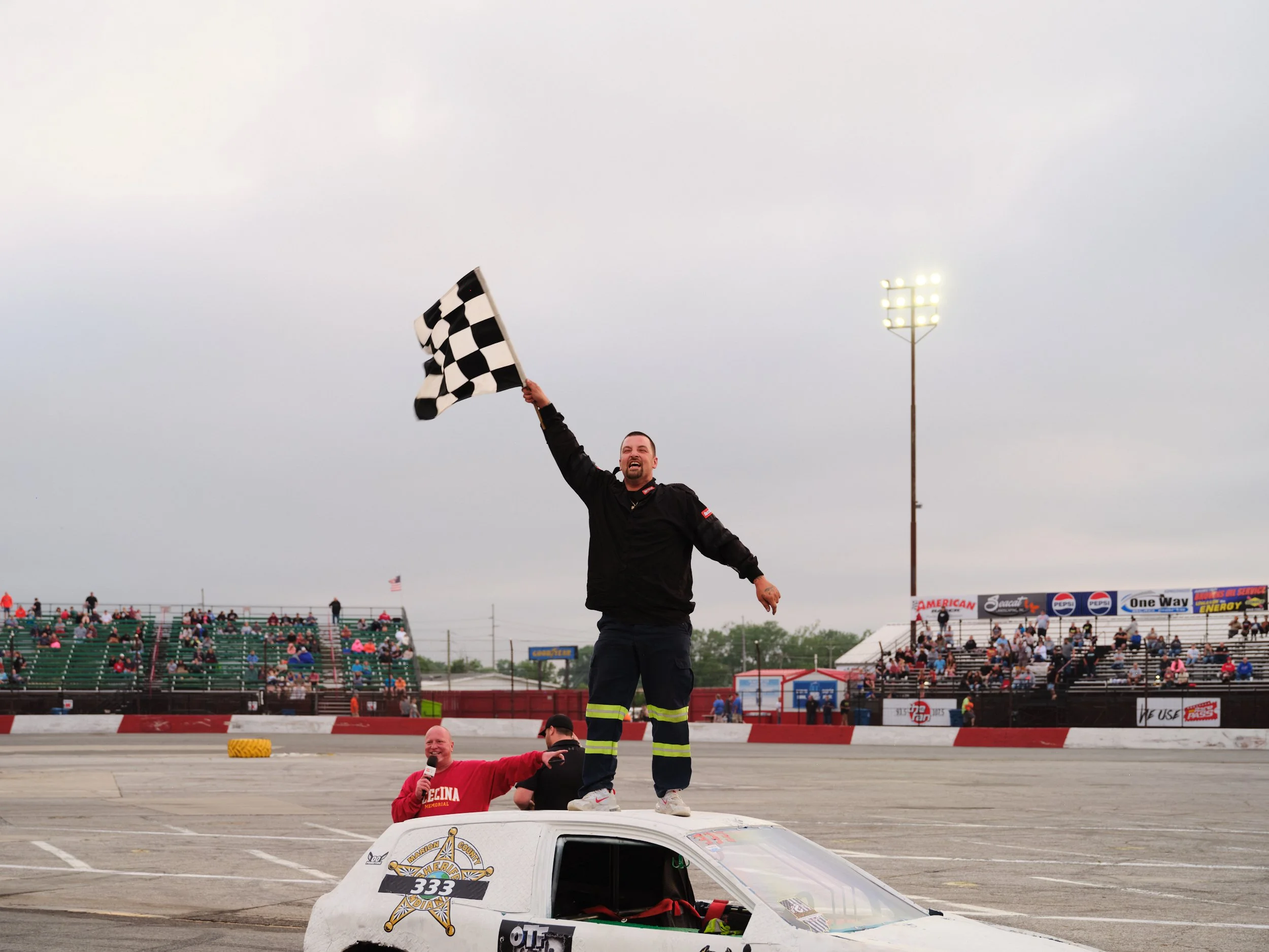 A man celebrating on top of a race car, holding a checkered flag, at a race track with spectators in the stands.