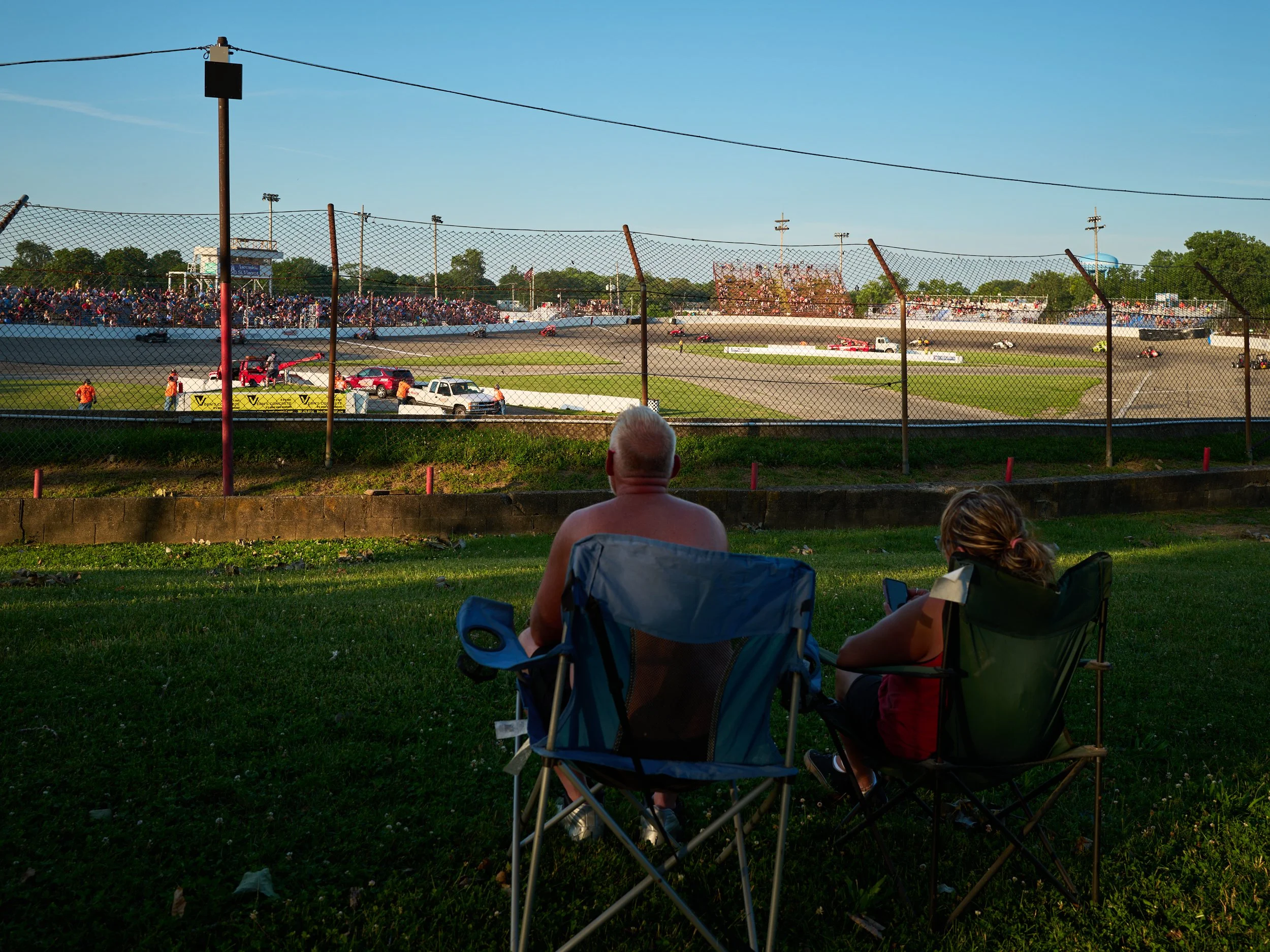 A man and a woman sitting in camping chairs on grass, watching a dirt race track with cars racing in the distance, behind a chain-link fence.
