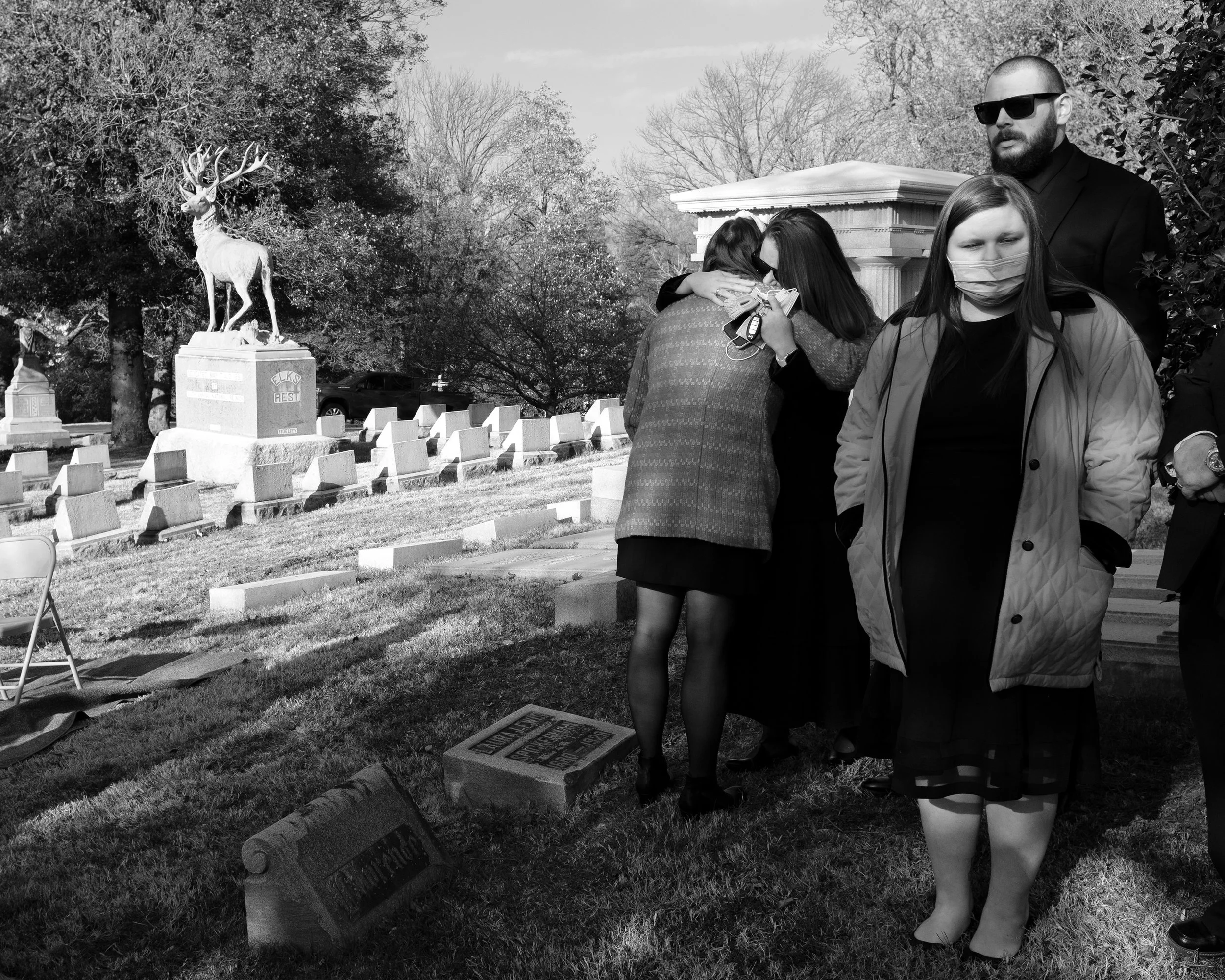 A group of people mourning at a gravesite with a statue of a deer in the background, some wearing masks, in a cemetery with tombstones and trees.