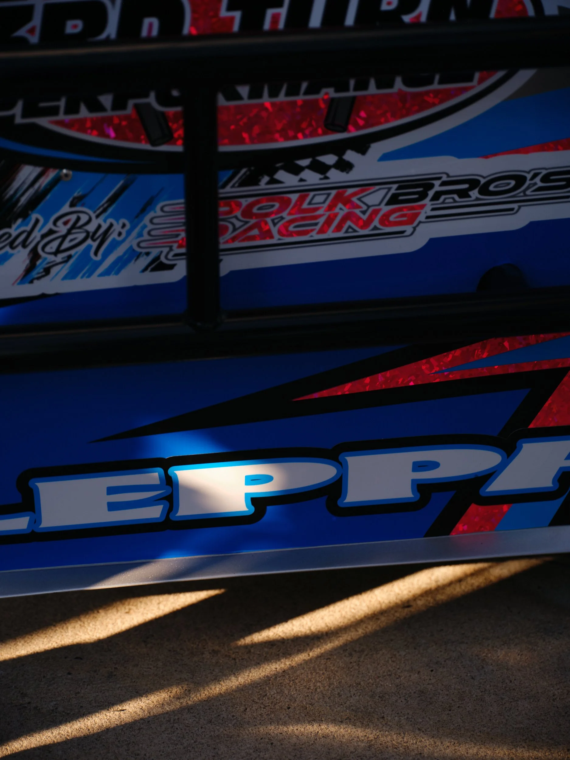 Close-up of a car with blue, red, and white decals, featuring the logos for EPPI and Falk Brothers Racing, and a protective cage over the window.