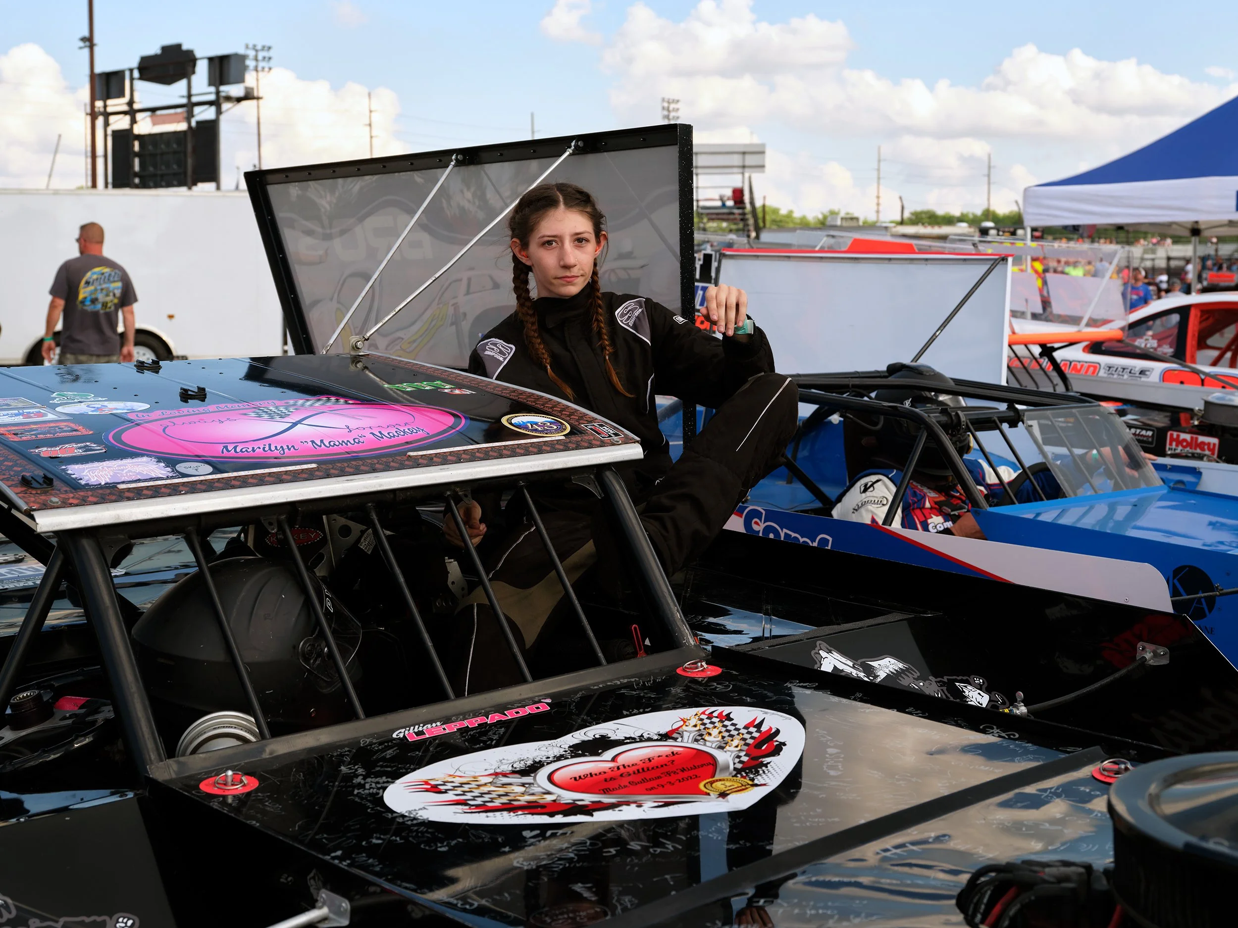 A young female race car driver with long braided hair, wearing a black racing suit, sitting on the side of a black race car with various stickers and decals, at a race track with other race cars and people in the background.