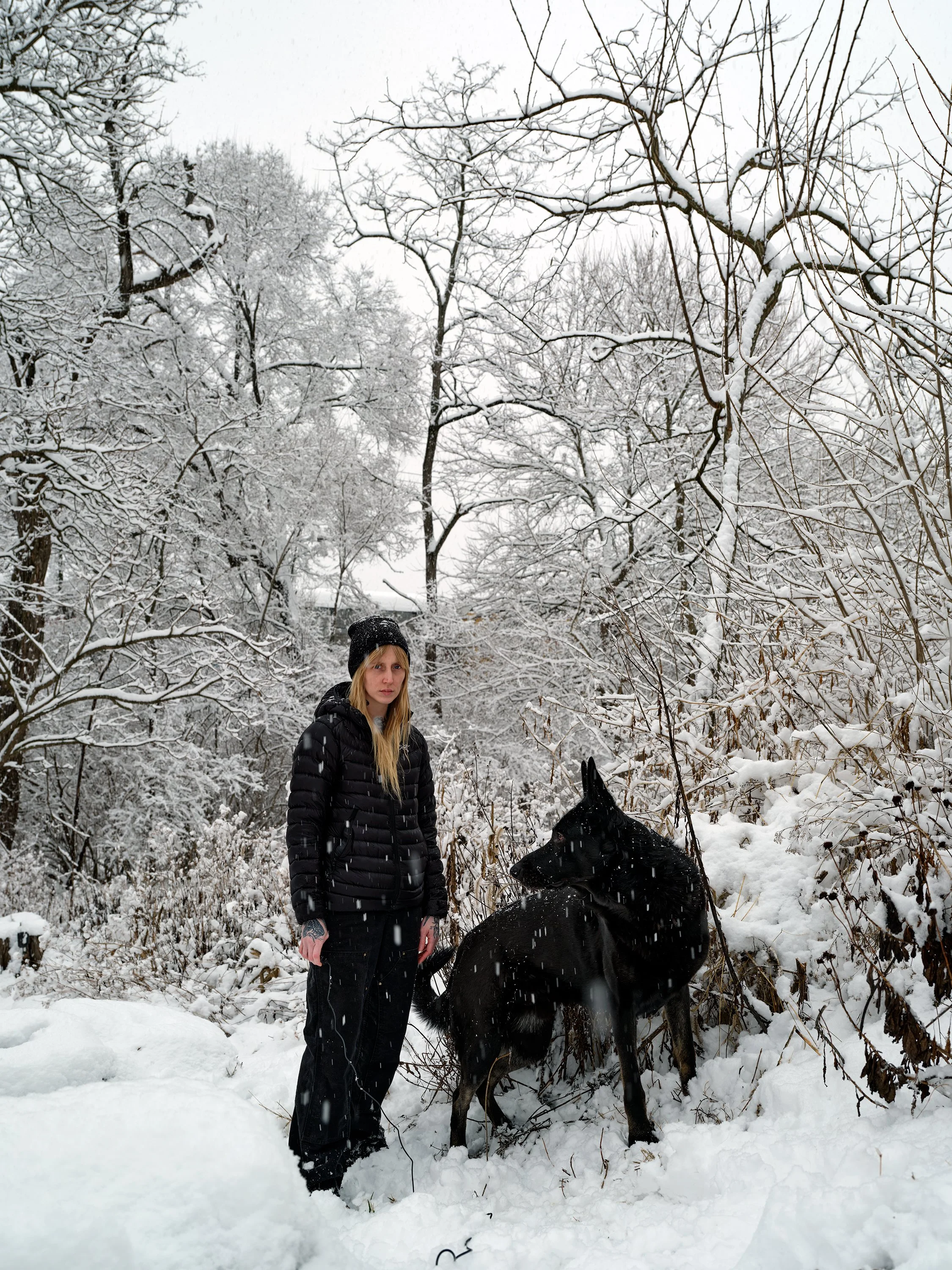 A woman dressed in black winter clothes standing in a snow-covered forest next to a black dog with tall ears, surrounded by snow-laden bare trees.
