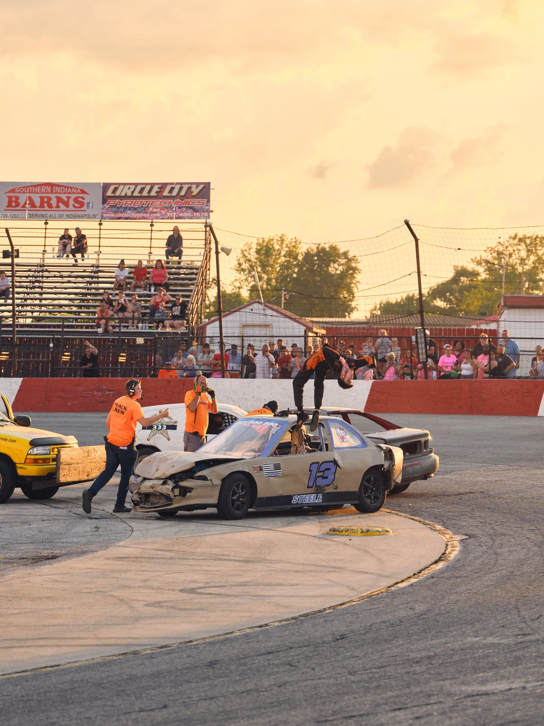 A damaged race car with the number 13 is stopped on a race track, with a person standing on top of it. Several crew members are working around the car, and spectators are sitting in the bleachers in the background, with a sunset sky overhead.