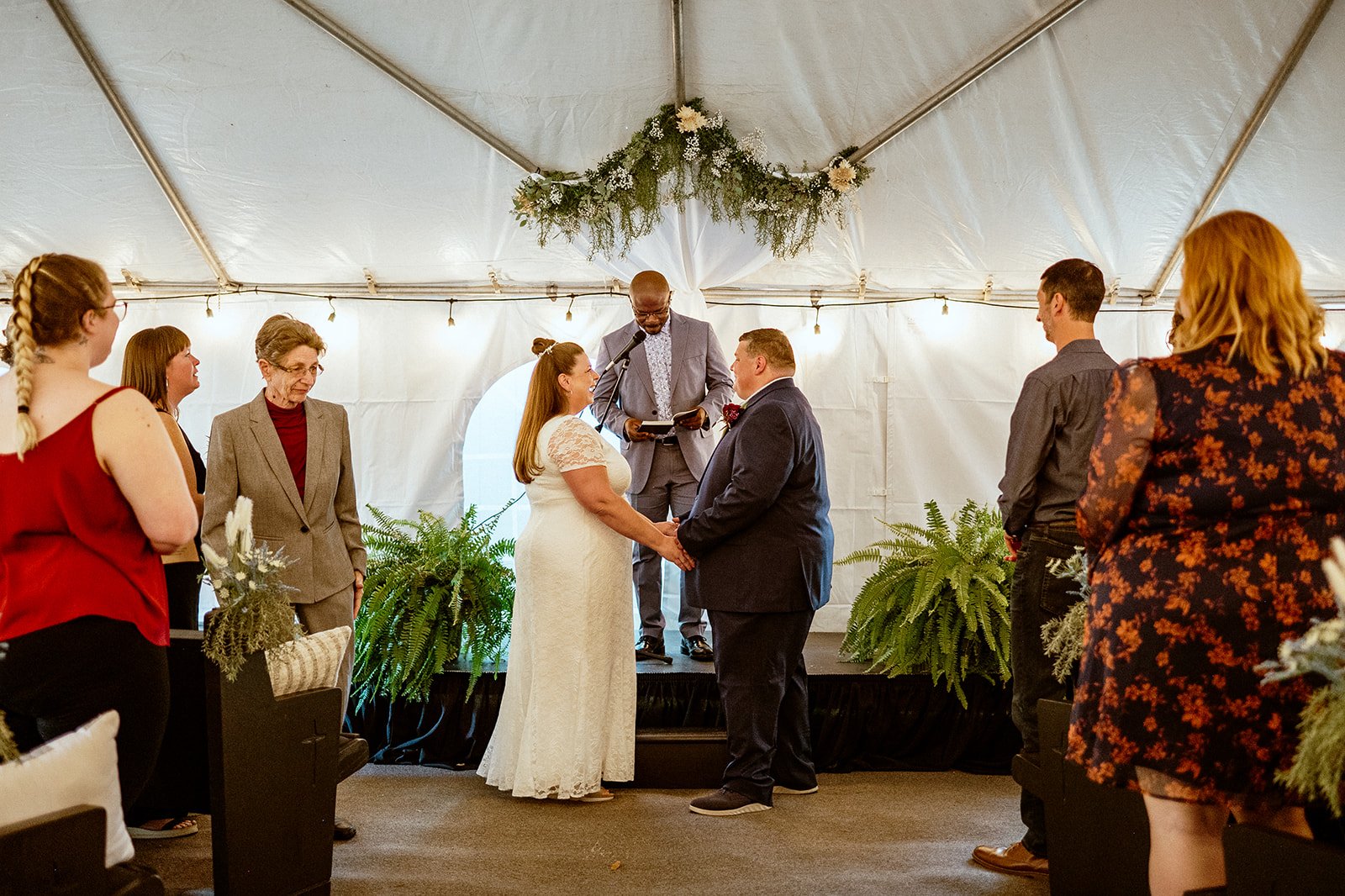 A wedding ceremony under a white tent with a bride and groom holding hands in front of an officiant. Guests stand on either side watching the couple. The tent is decorated with flowers and greenery.