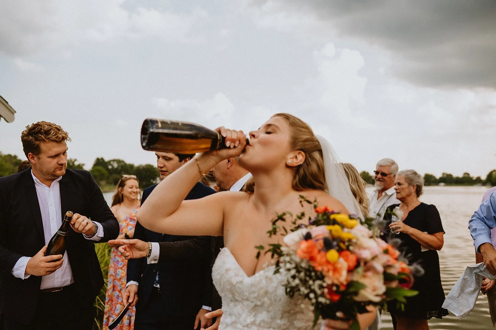Bride drinking from a wine bottle while holding a bouquet, surrounded by guests at a lakeside event.