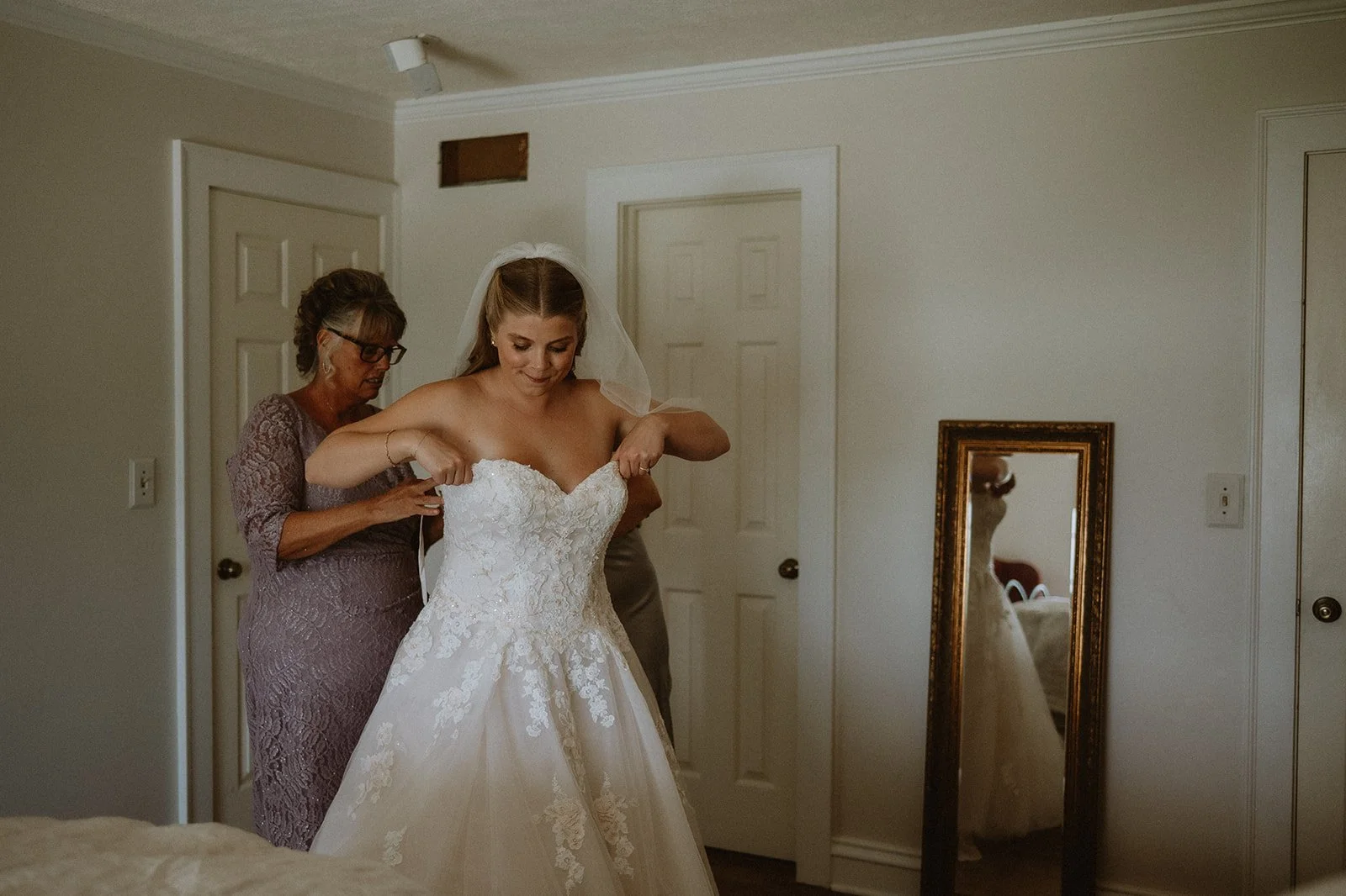 Bride adjusting wedding dress helped by older woman in bedroom, mirror reflecting gown.