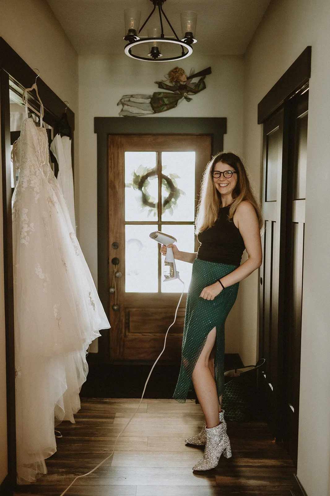 Woman steaming a white lace wedding dress in a hallway with wooden floors, wearing a black top, green skirt, and sparkly boots.