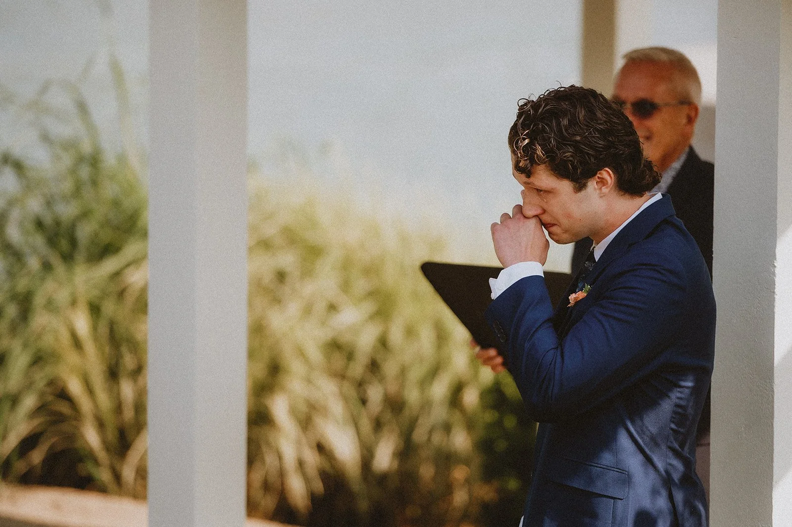 Man in a blue suit standing outdoors with emotional expression, holding his hand to his face next to a man in sunglasses holding a folder.