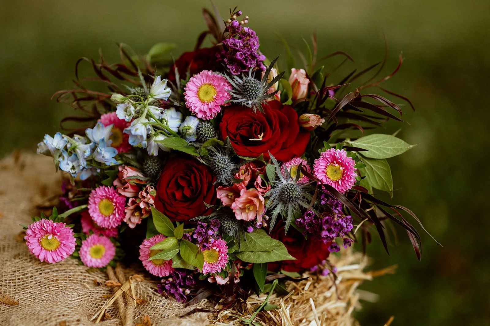 Colorful floral bouquet with red roses, pink asters, blue delphiniums, and greenery on burlap.
