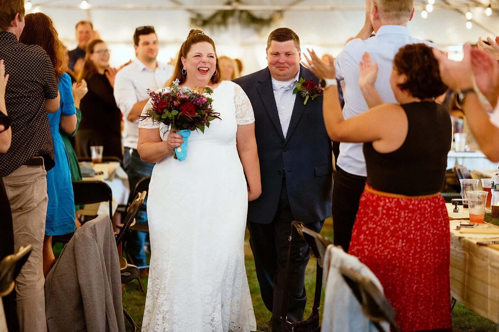 Bride and groom smiling and walking down the aisle at a wedding reception, surrounded by applauding guests under a tent.