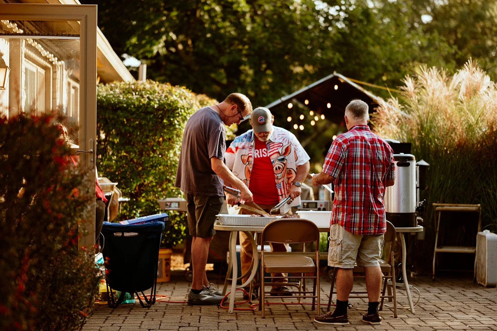Three men preparing food at an outdoor barbecue event, surrounded by greenery. One man wears a colorful shirt with a pig design, while another wears a red plaid shirt. They are using cooking utensils over a table with folding chairs nearby. String li
