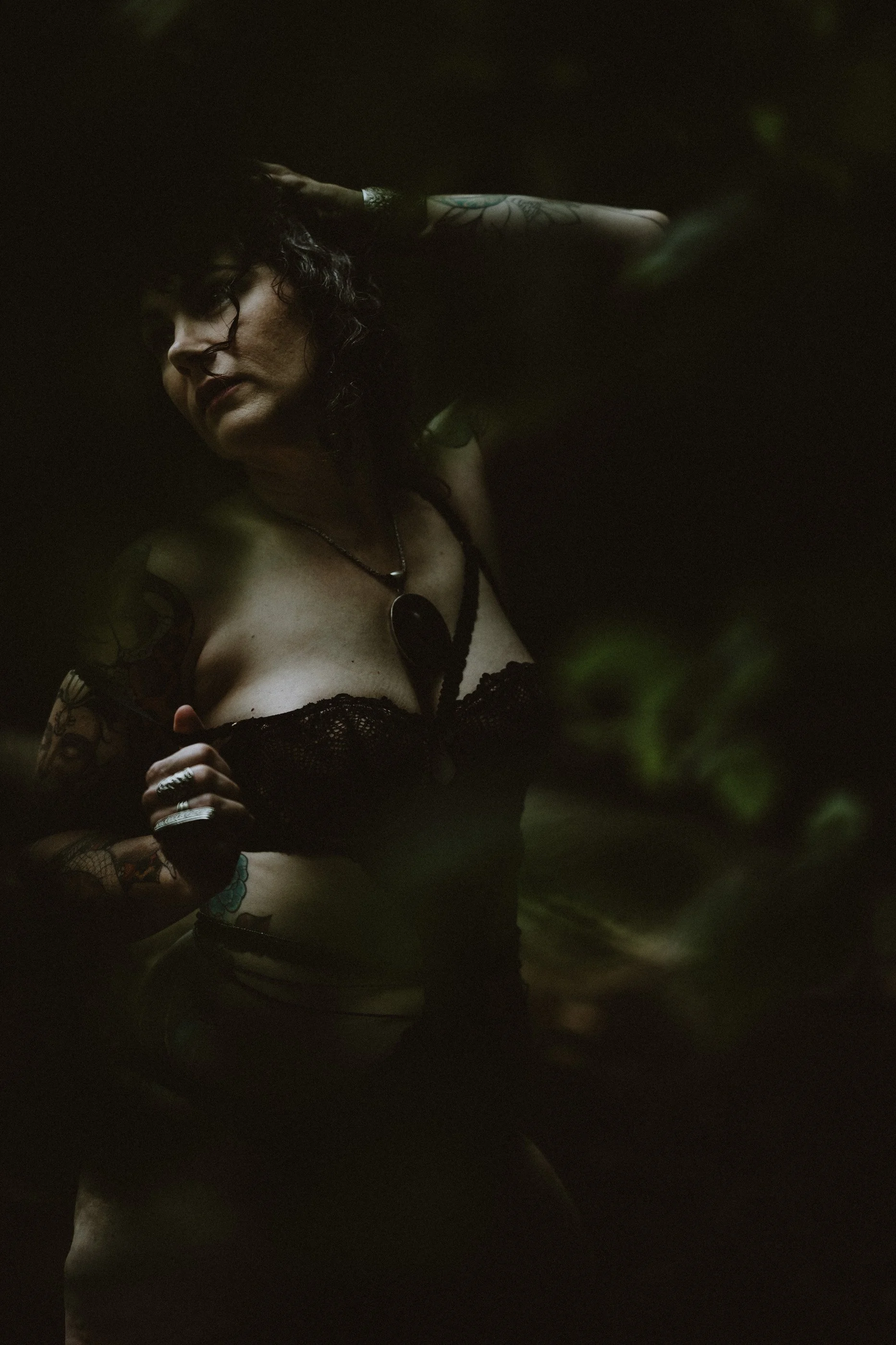 A woman in dark lingerie poses amid shadowy foliage.