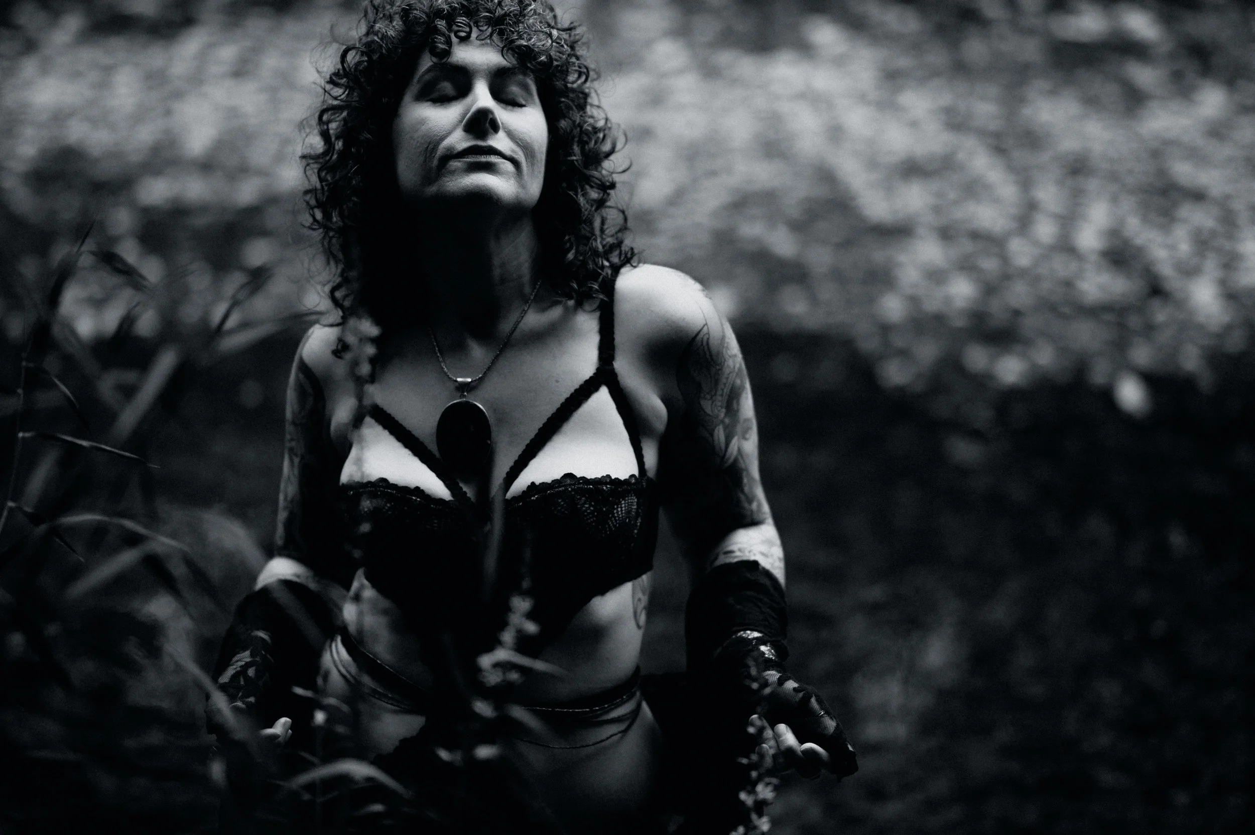 Black and white photo of a person with curly hair in a lace top, standing outdoors with eyes closed.