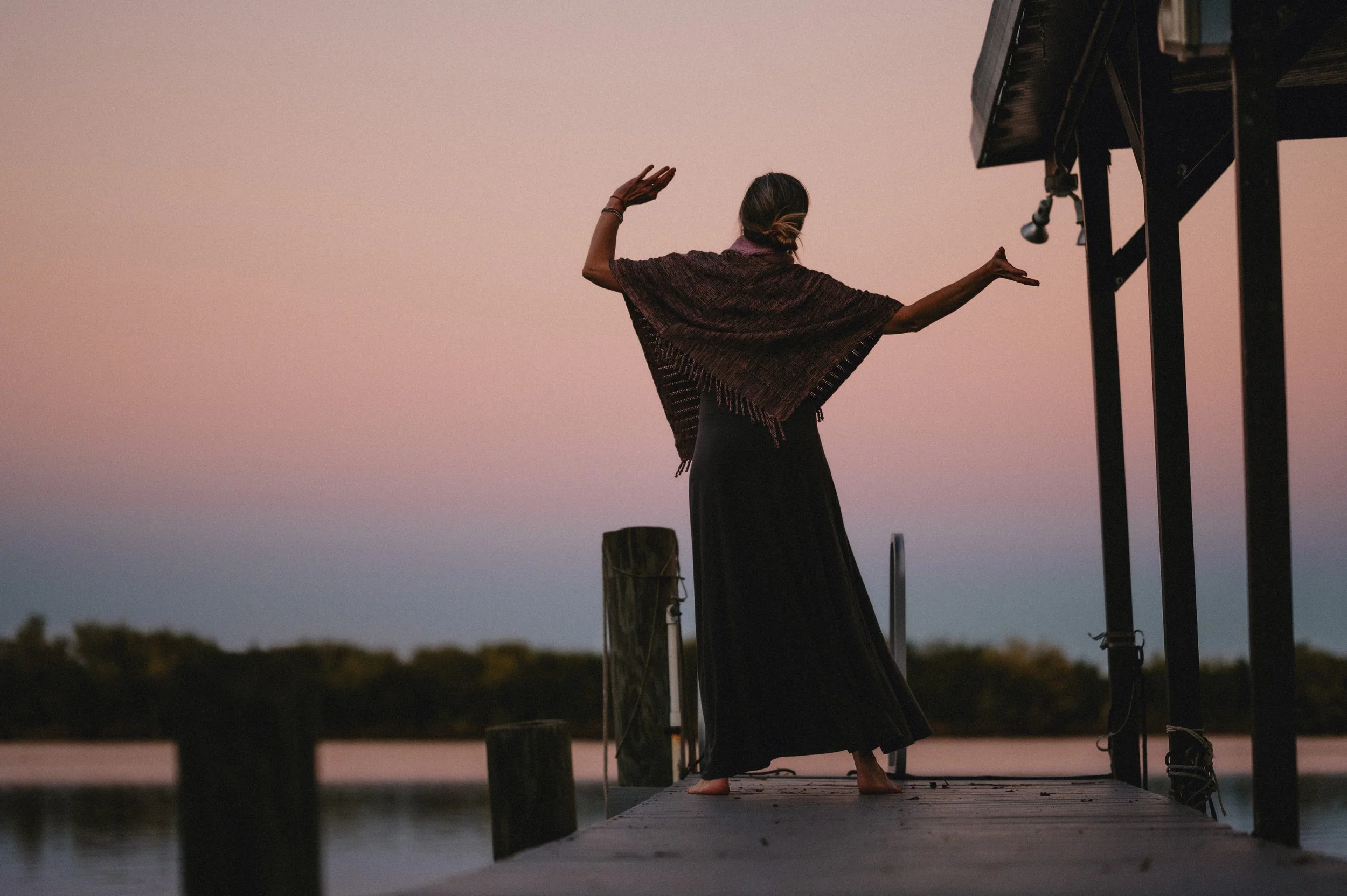 Silhouette of a person dancing on a dock at sunset with a shawl, near a calm lake and trees in the background.