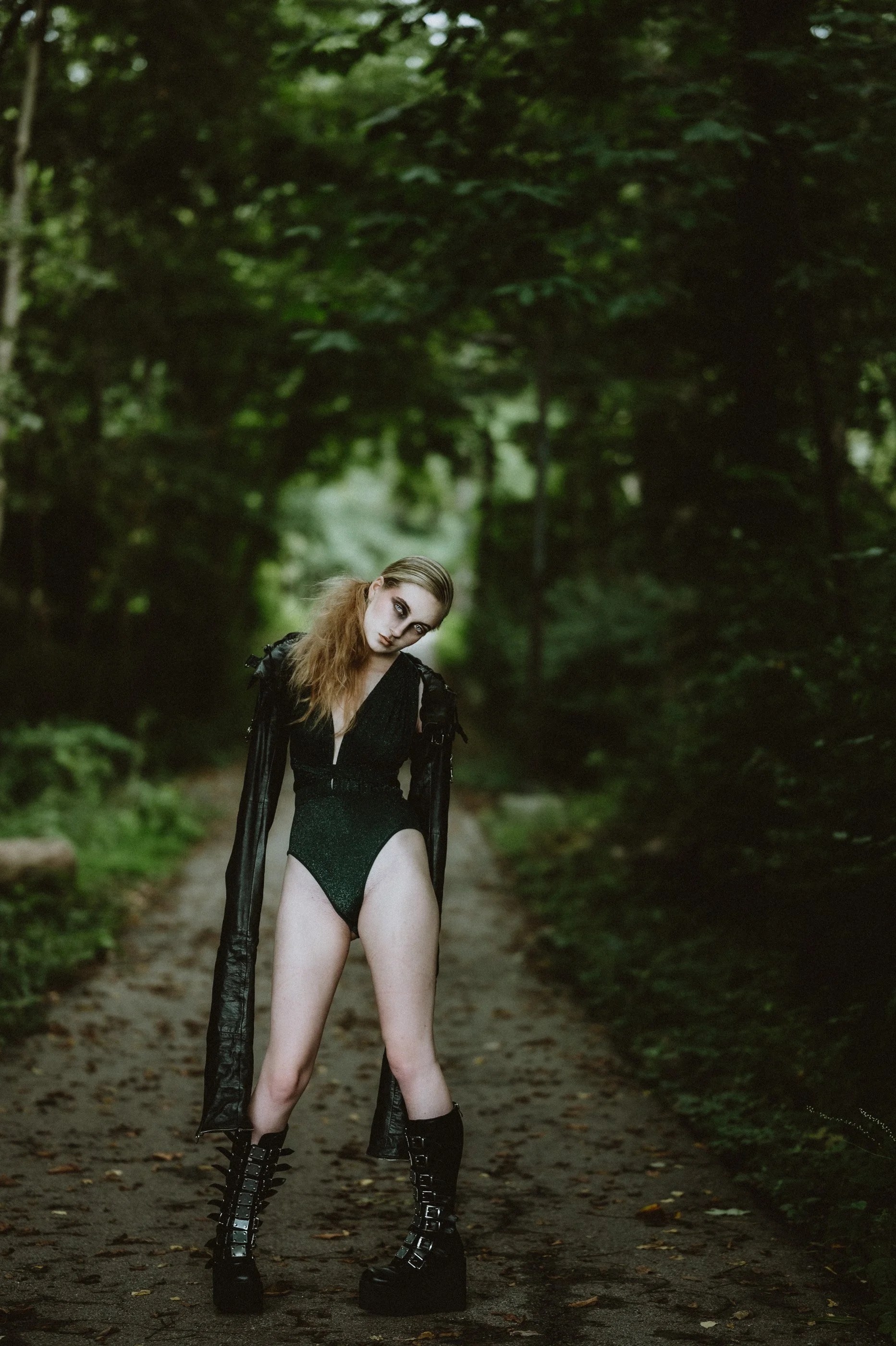 A person wearing a black bodysuit, long leather coat, and platform boots stands on a forest path.