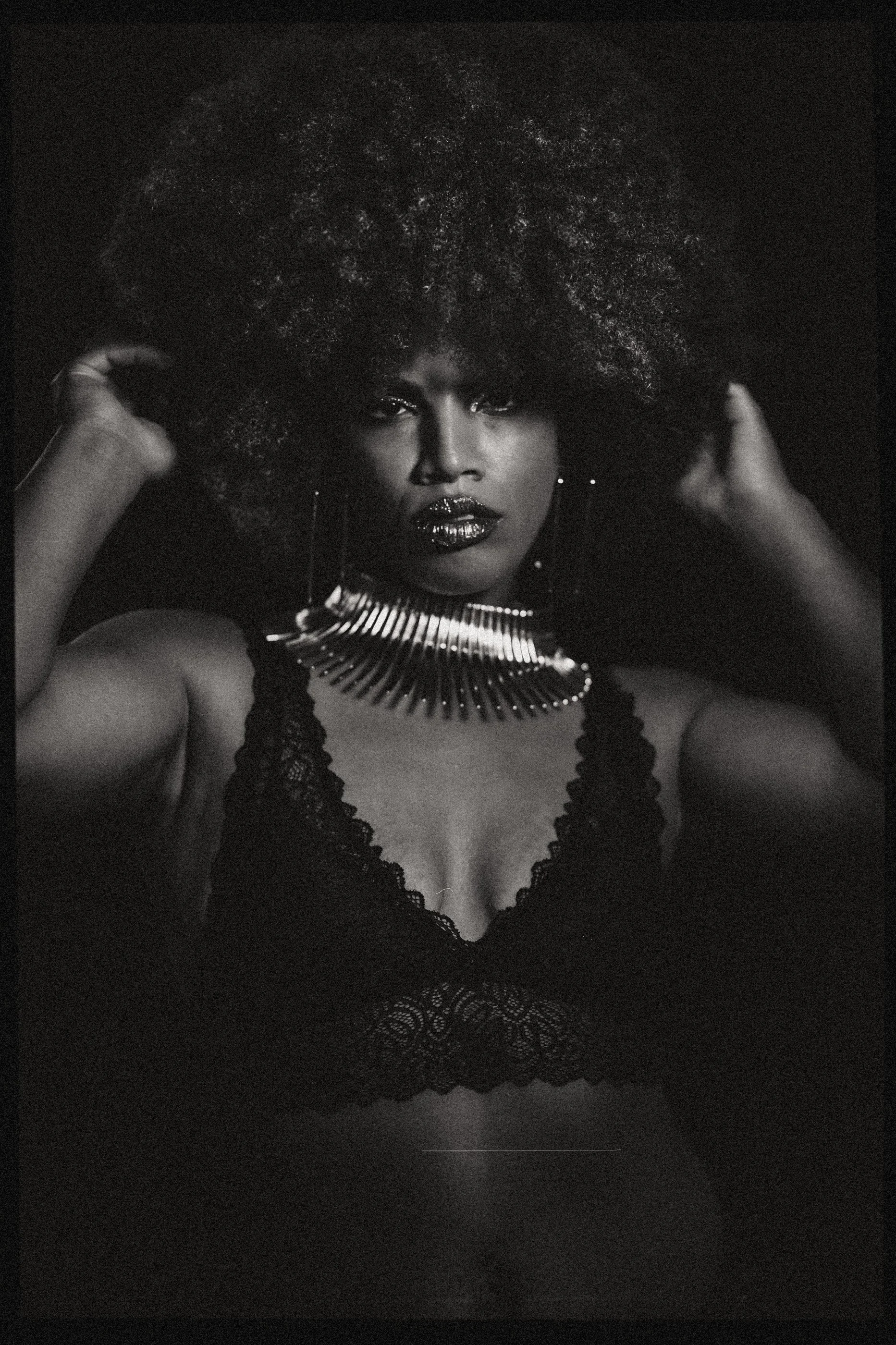 Black and white portrait of a person with an afro hairstyle, wearing a lace top and statement necklace.