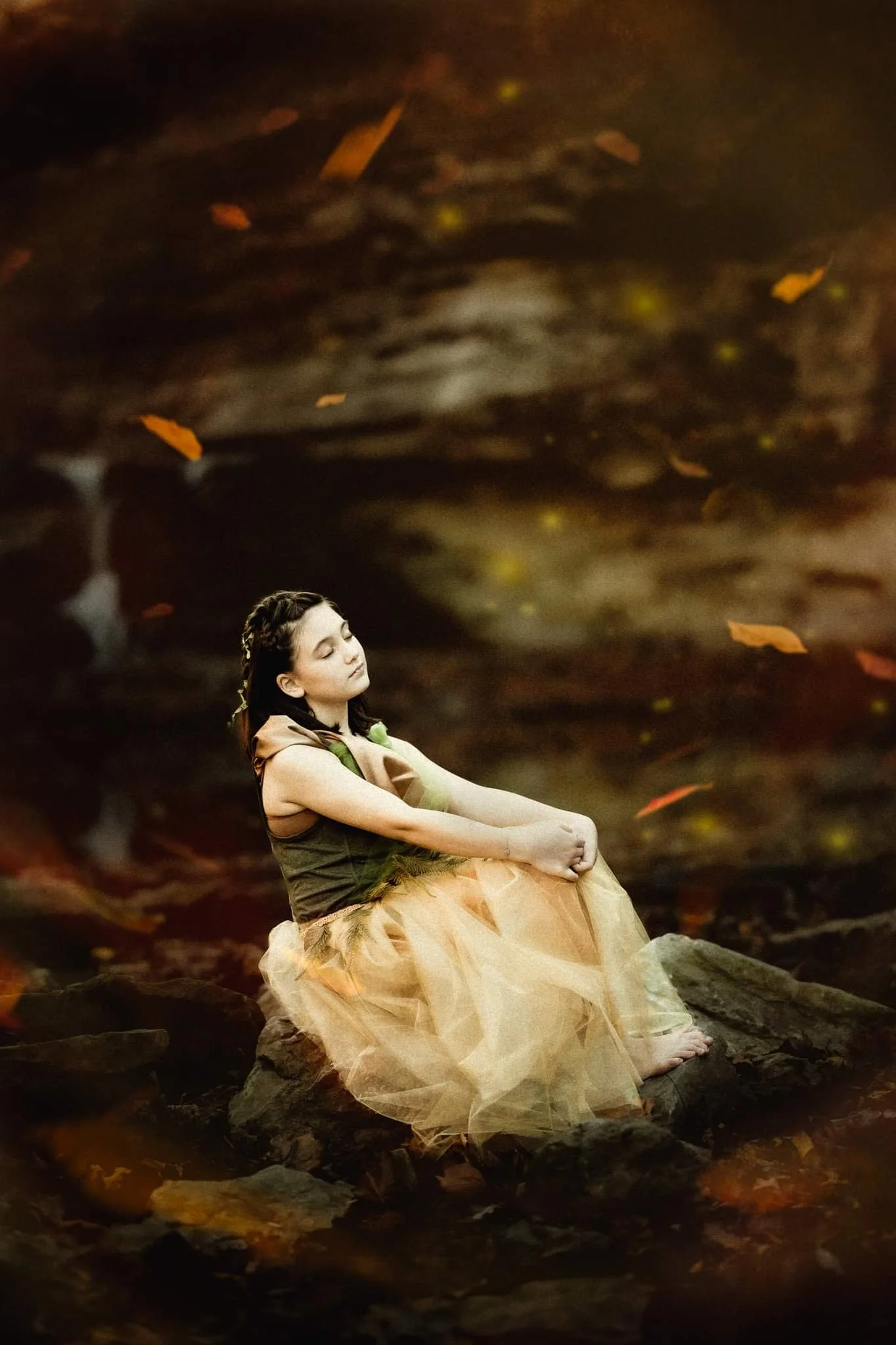 Young girl in a dress sitting on rocks with a dreamy expression, surrounded by autumn leaves.