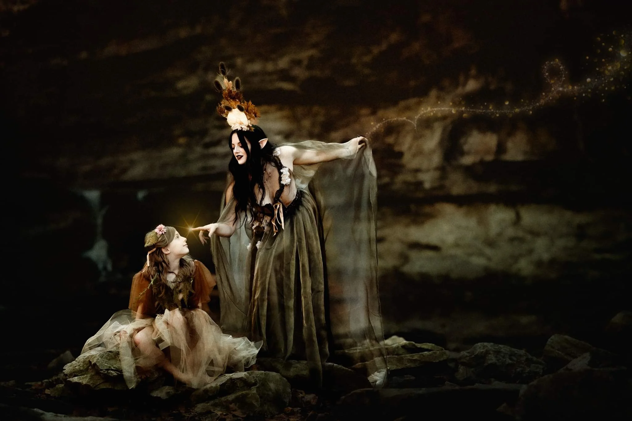 A mystical scene with two women dressed as fantasy characters; one standing and one sitting on rocks in an enchanted forest setting. The standing woman extends her hand toward the other, creating a magical light and sparkles.