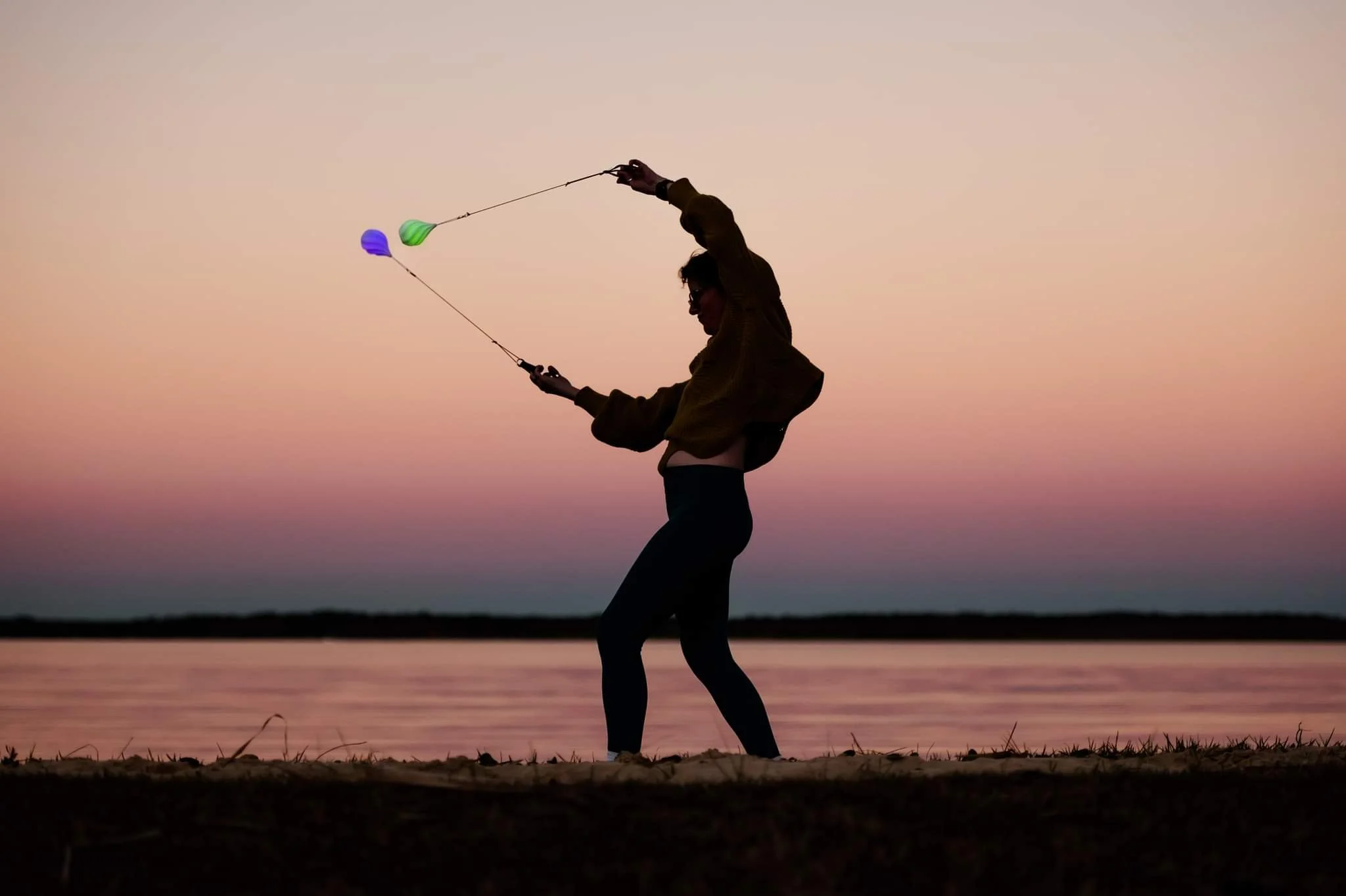 Silhouette of a person spinning LED poi near a body of water at sunset.