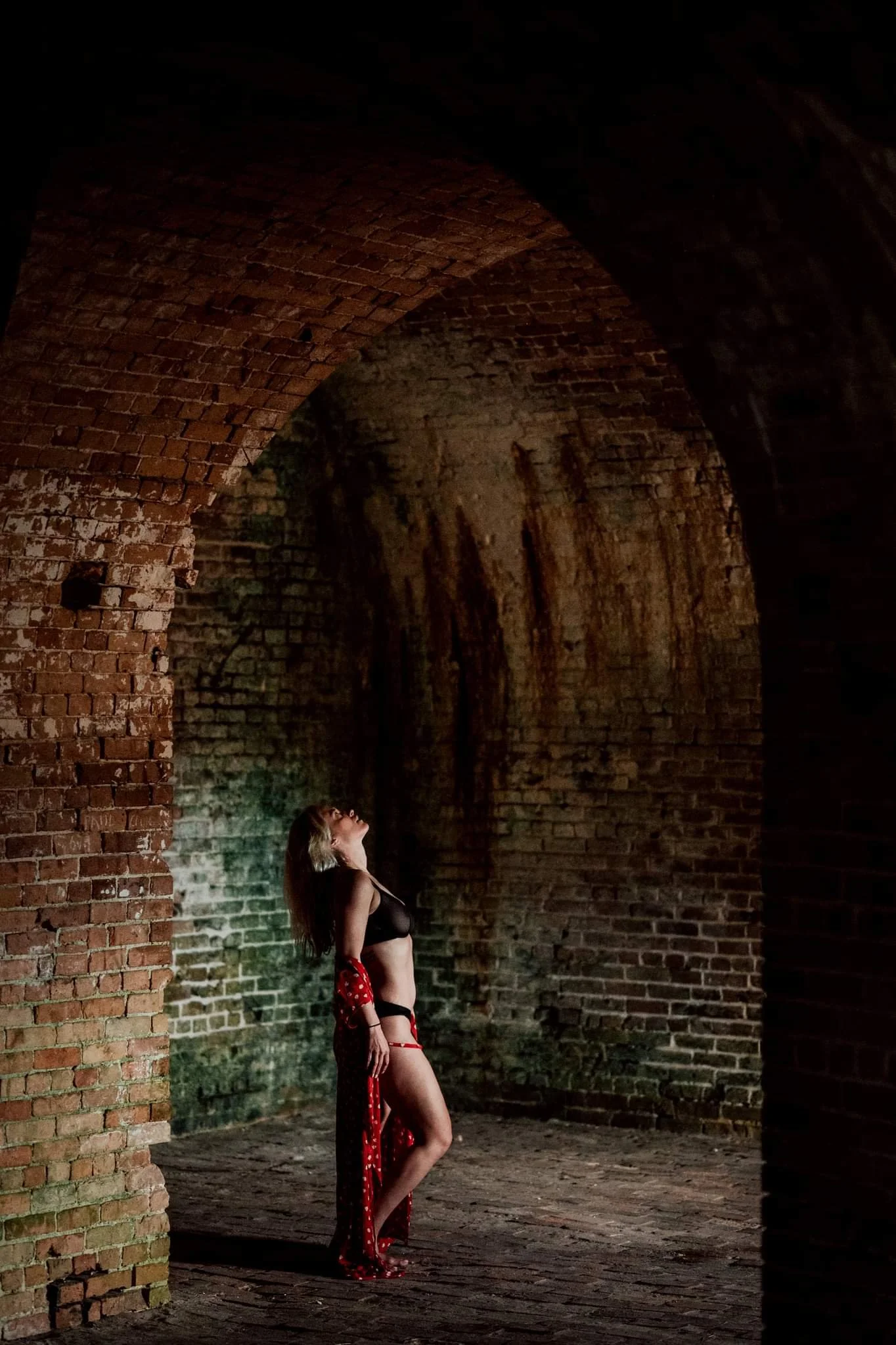 A person in lingerie poses dramatically under a brick archway in a dimly lit space with textured brick walls.