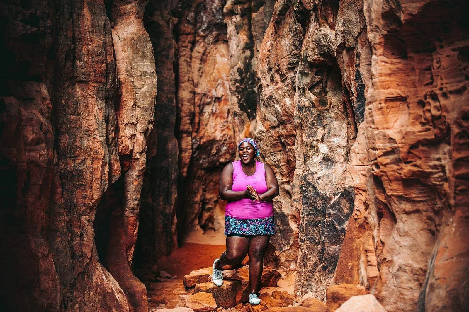 Woman in pink tank top standing between large red rocks, smiling.