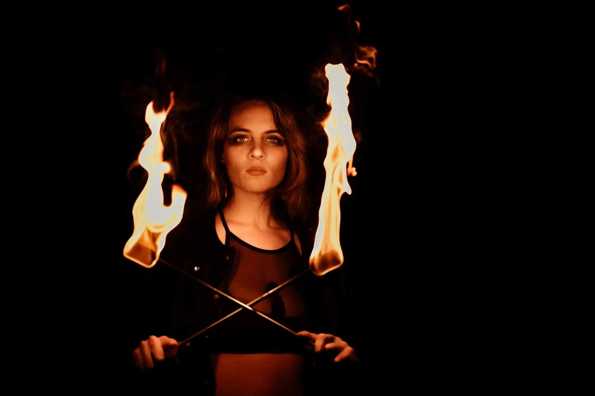 Person holding two lit fire torches crossed in front of their body against a dark background.