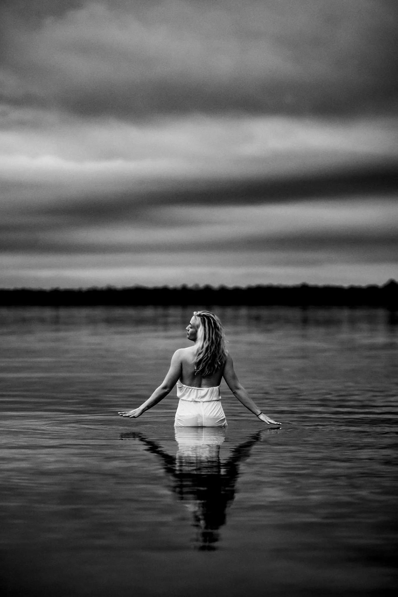 A woman in a white outfit standing in shallow water, facing away, under a cloudy sky.