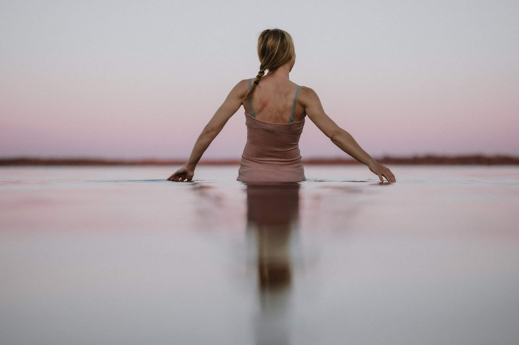 Woman sitting in water at sunset with a calm pink sky.