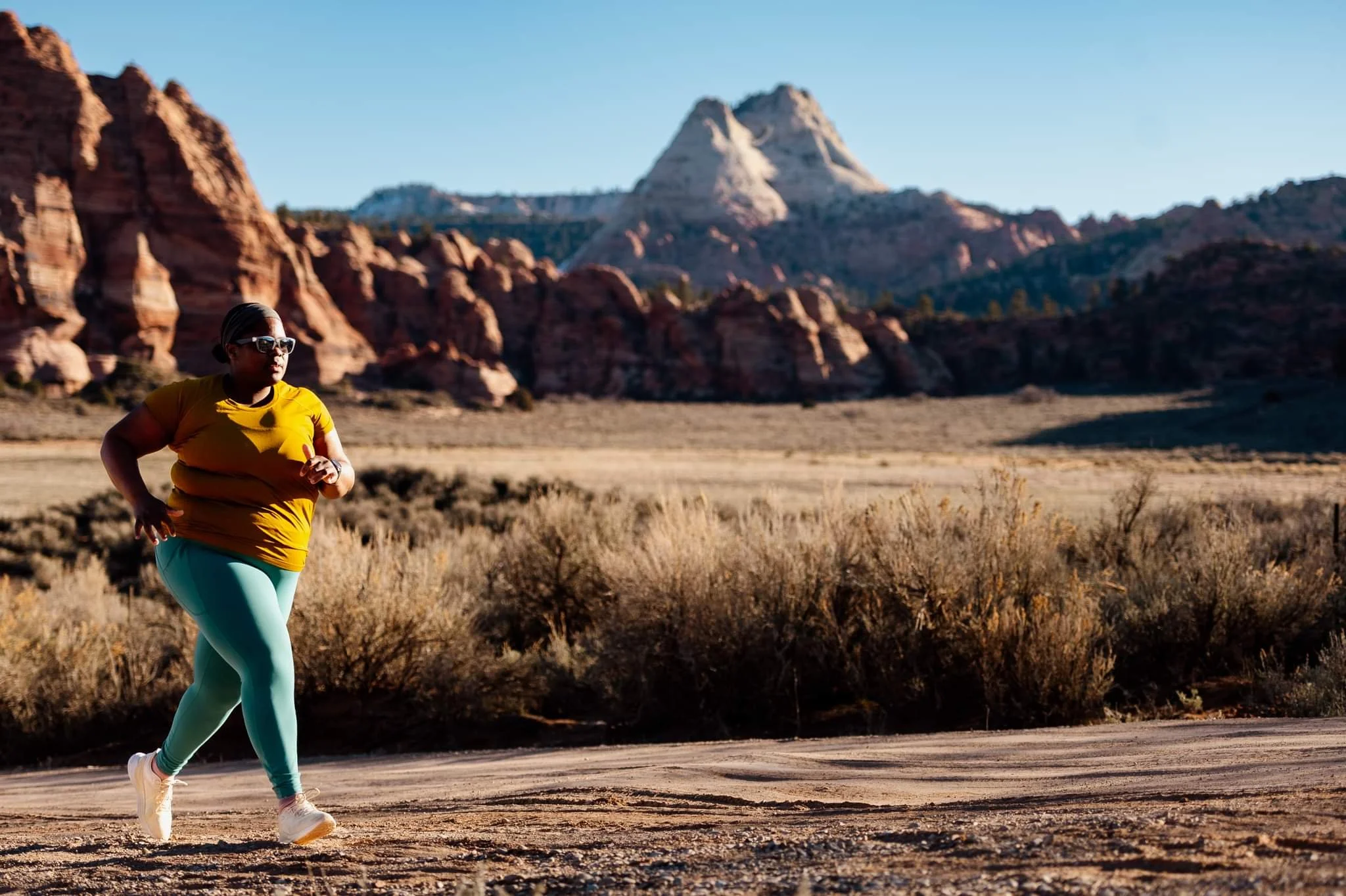 Person jogging on a trail in a desert landscape with mountains and clear blue sky.