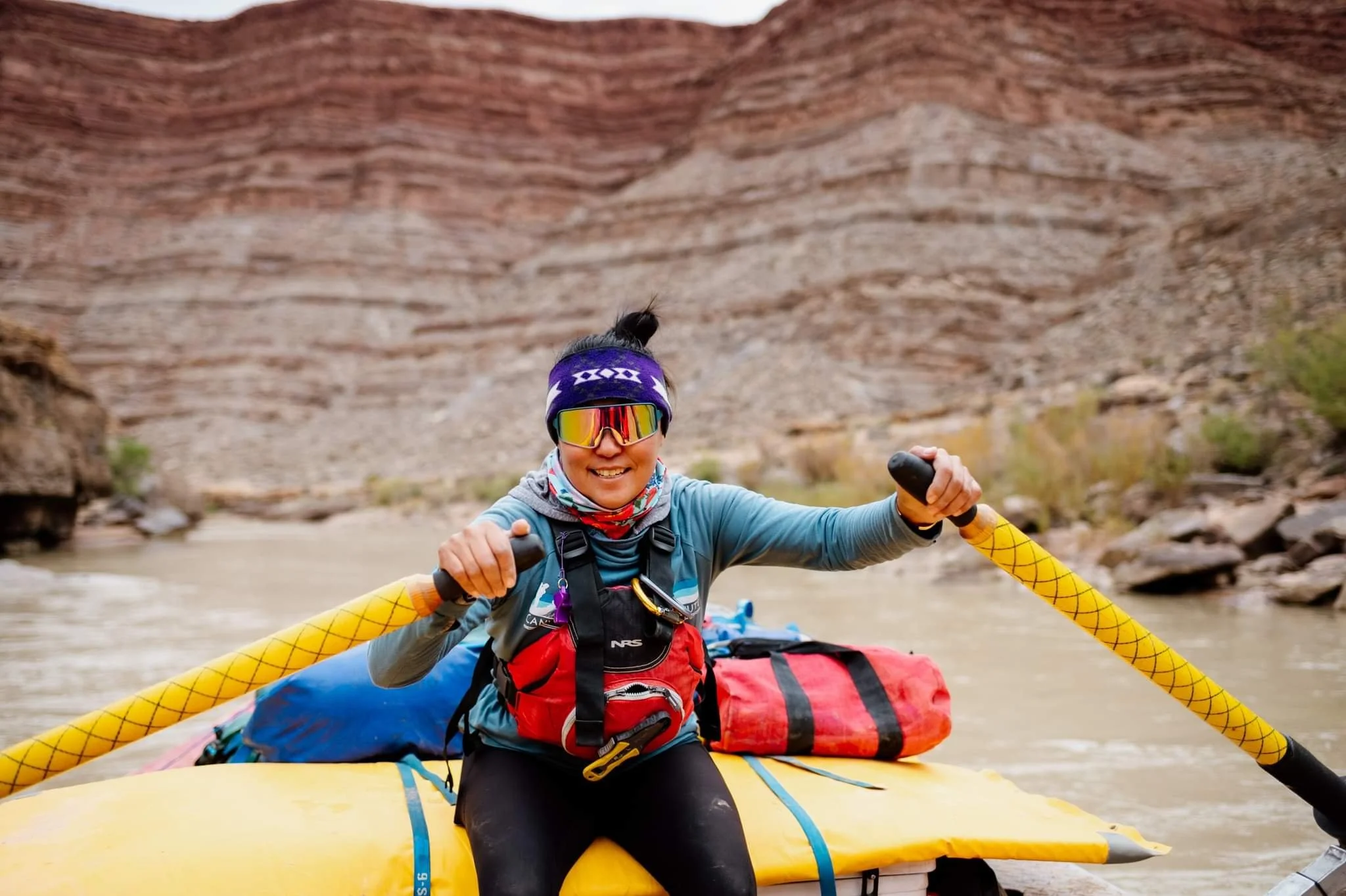 Person rafting on a river surrounded by rocky cliffs, wearing sunglasses and a red life jacket.