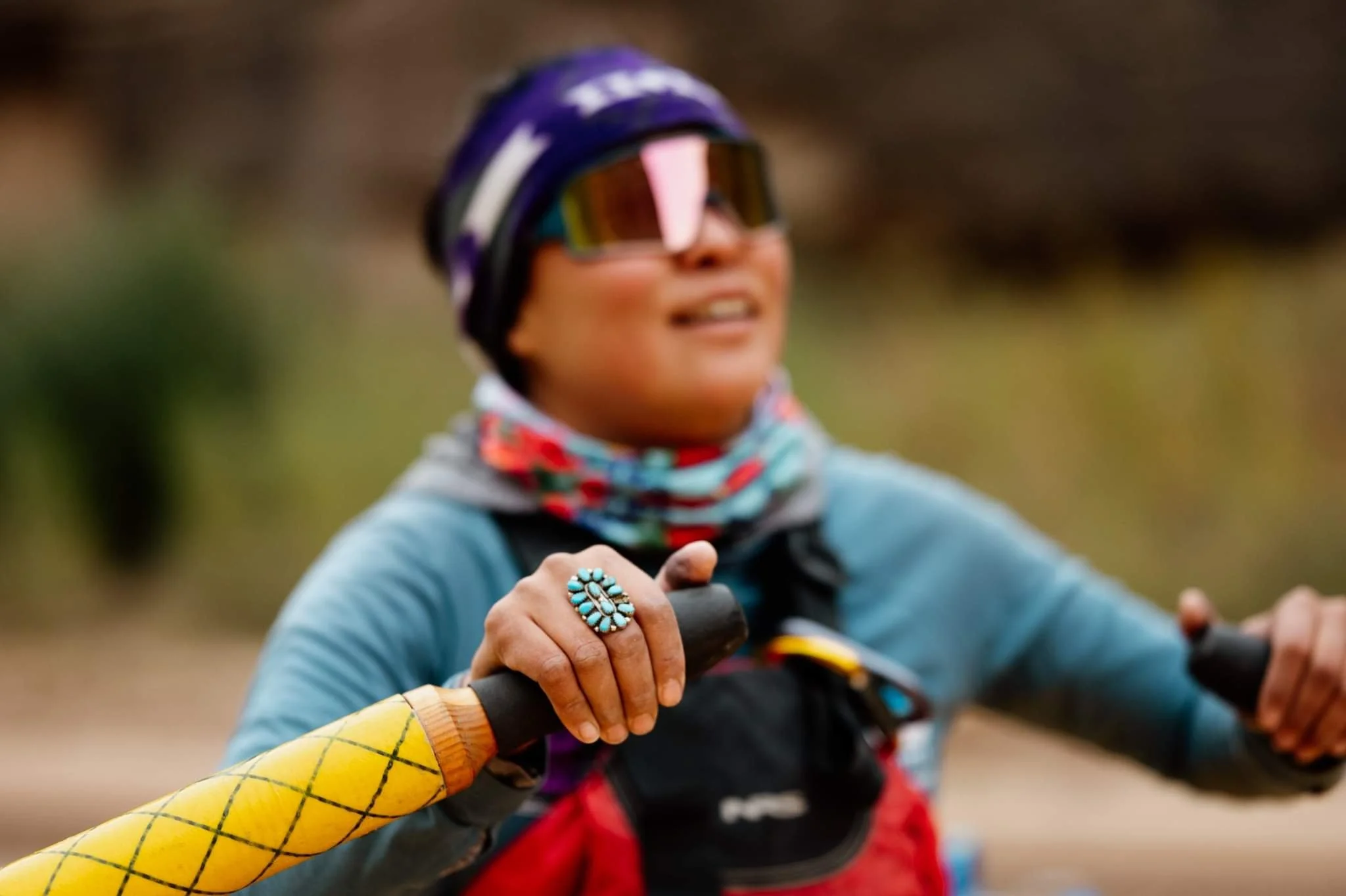 Person rowing with a yellow-handled oar, wearing sunglasses, a colorful scarf, and a beanie, featuring a turquoise ring on their finger.