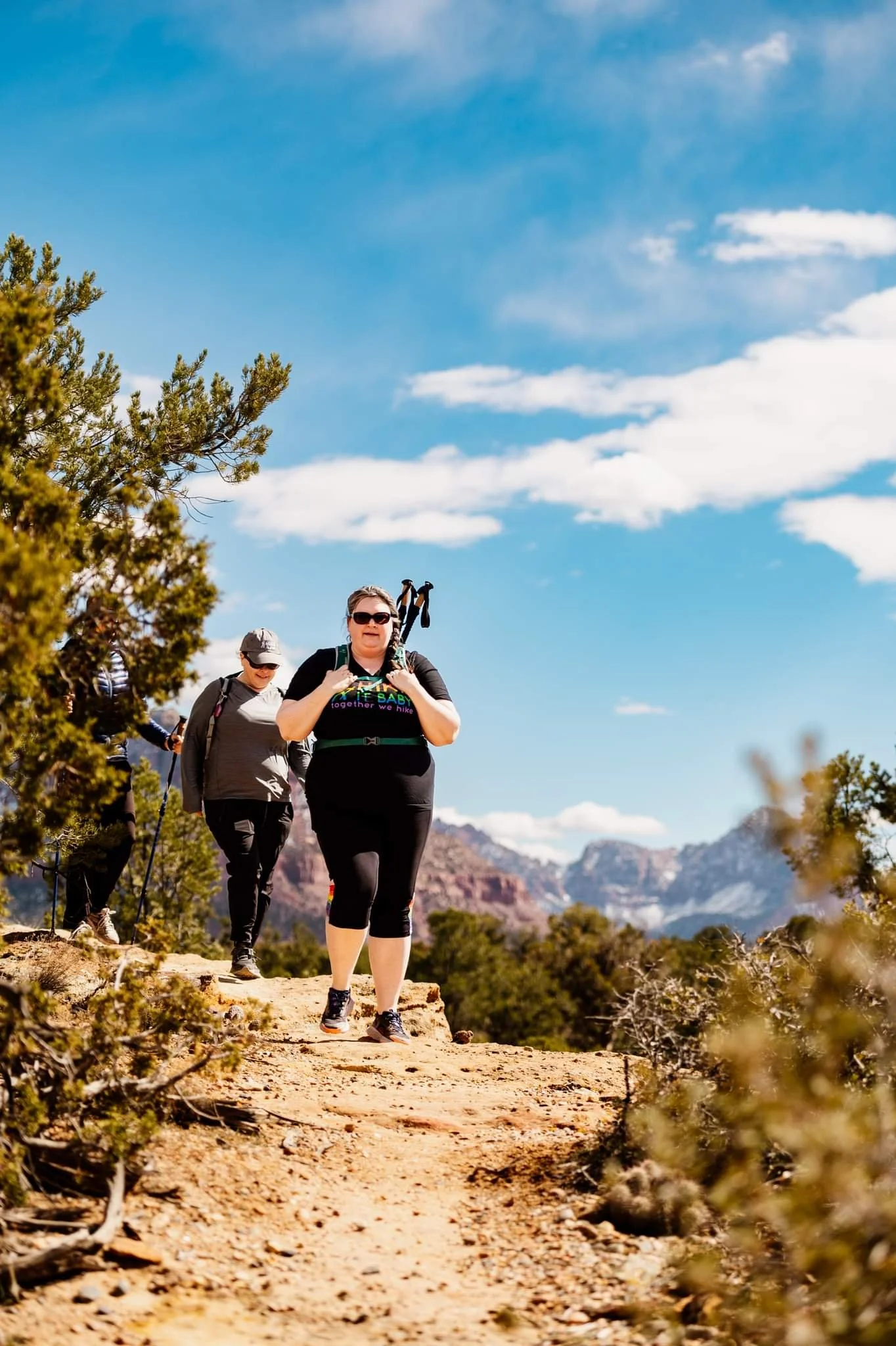 Three people hiking on a rocky trail with mountains in the background and clear blue sky.