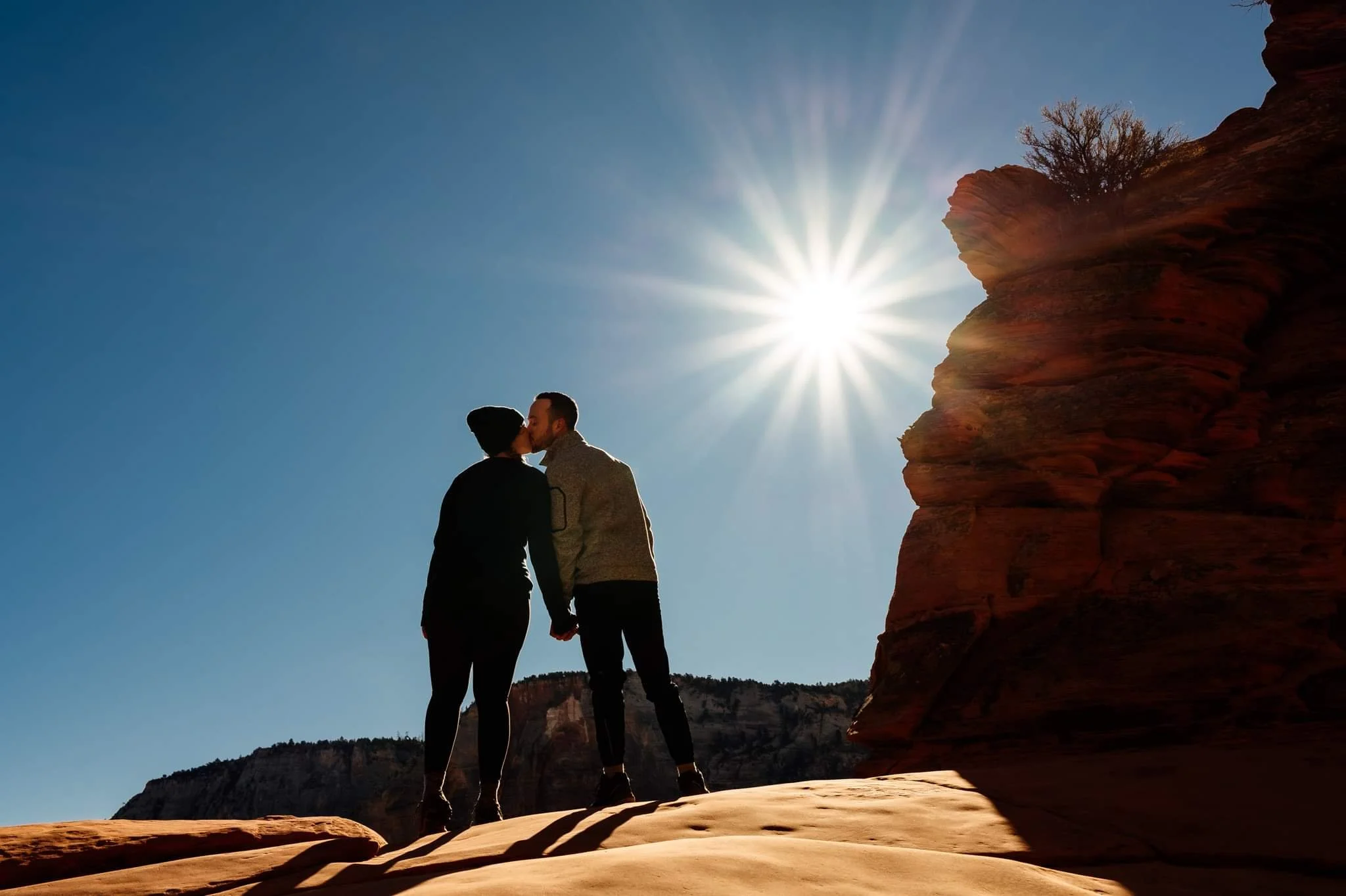 Couple kissing on sunlit rock formation in desert landscape.