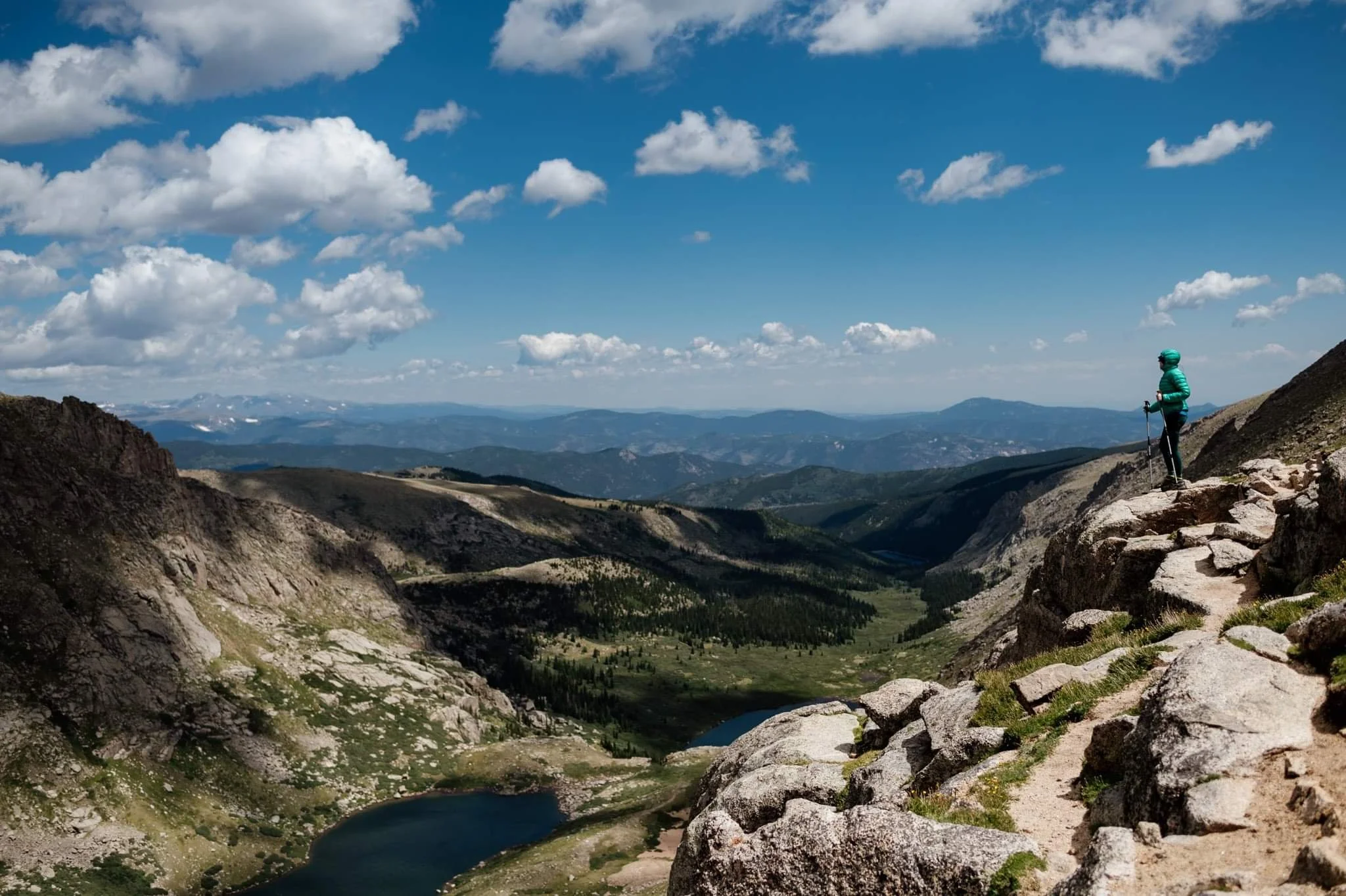 Hiker standing on rocky mountain trail with trekking poles, overlooking scenic valley, lakes, and mountains under a cloudy blue sky.