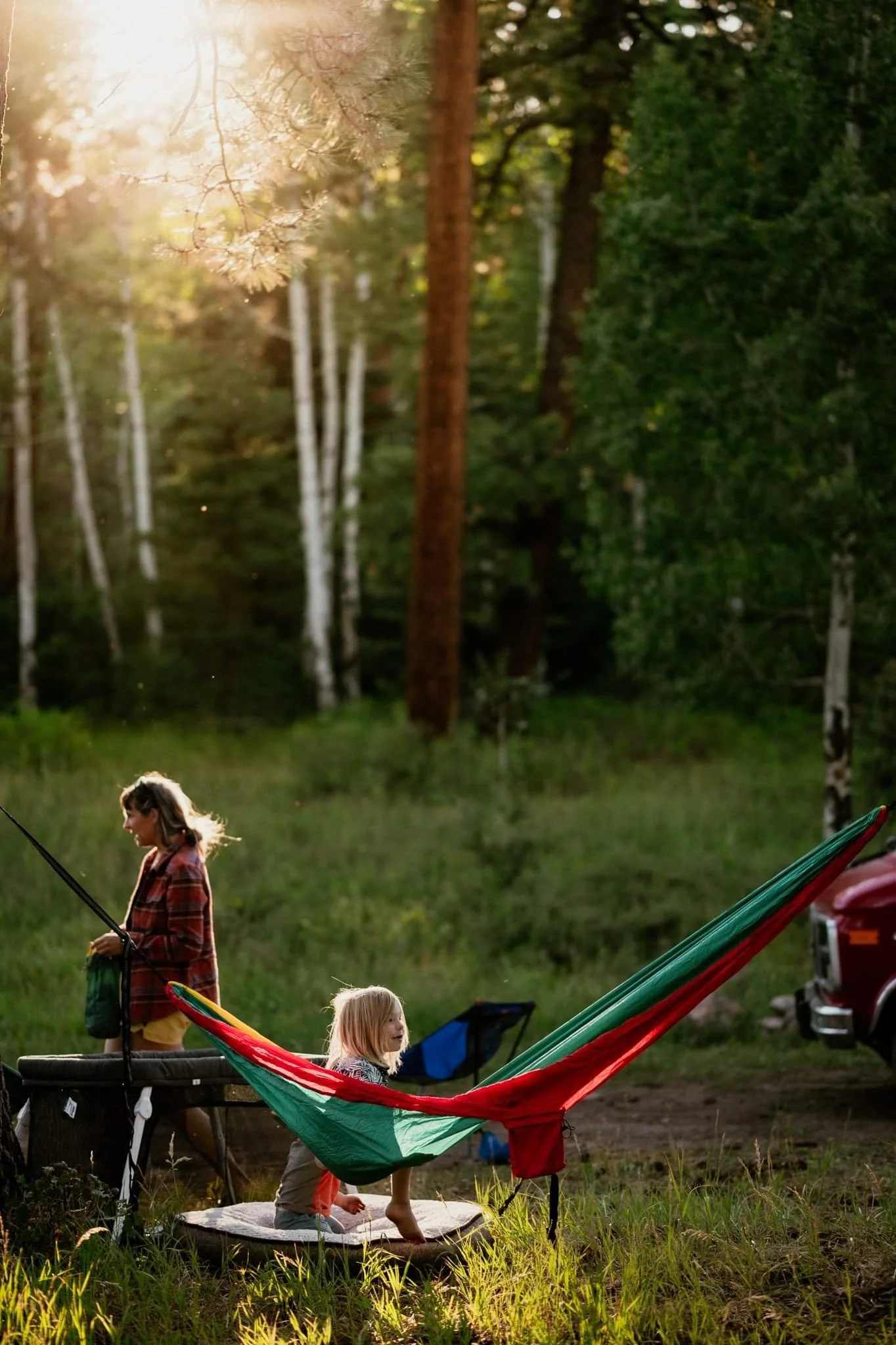 Child sitting in a hammock in a forest setting with a woman walking nearby and a car parked in the background under the sunlight.