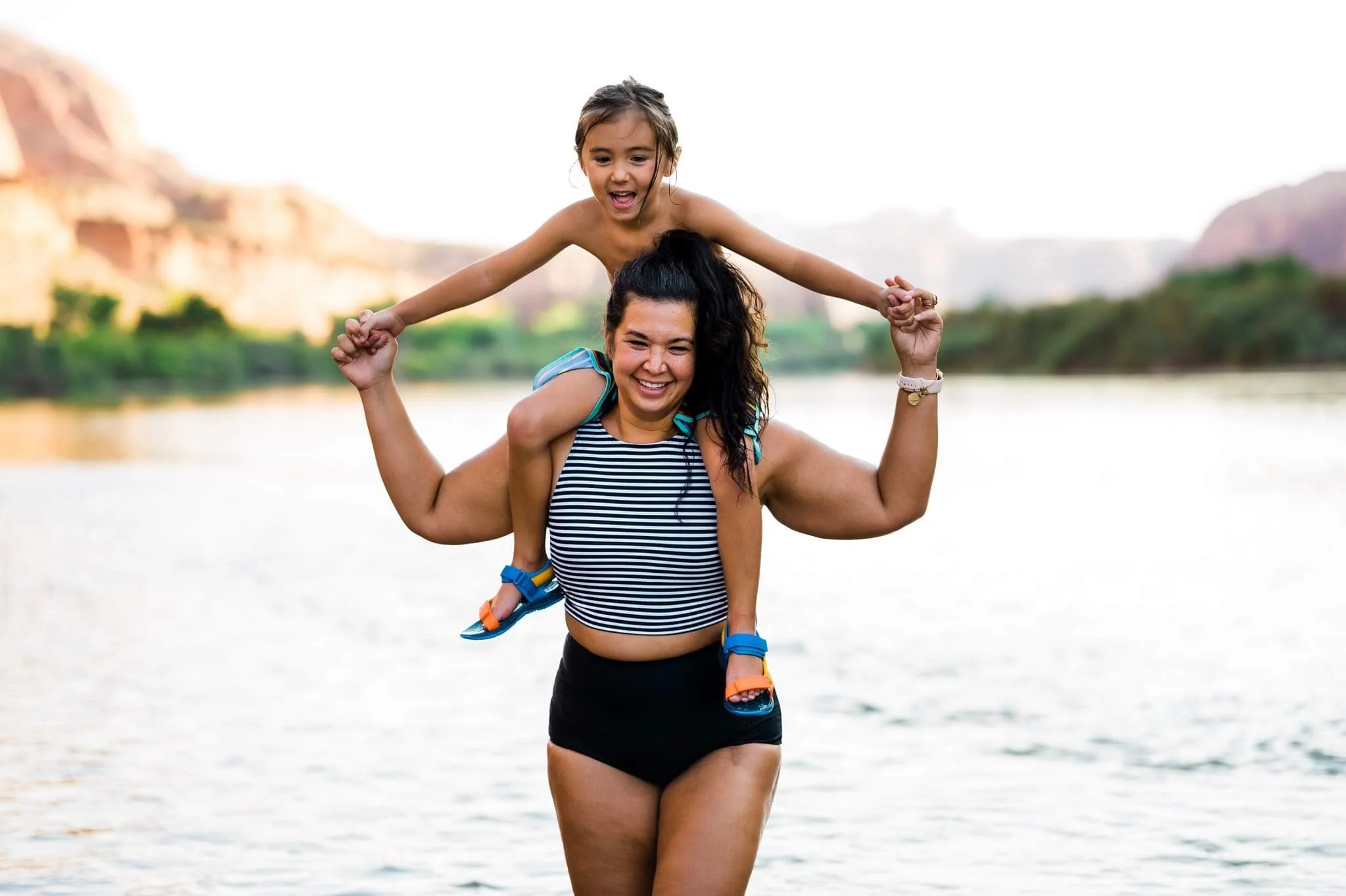 Woman smiling with child on shoulders near a river in nature.