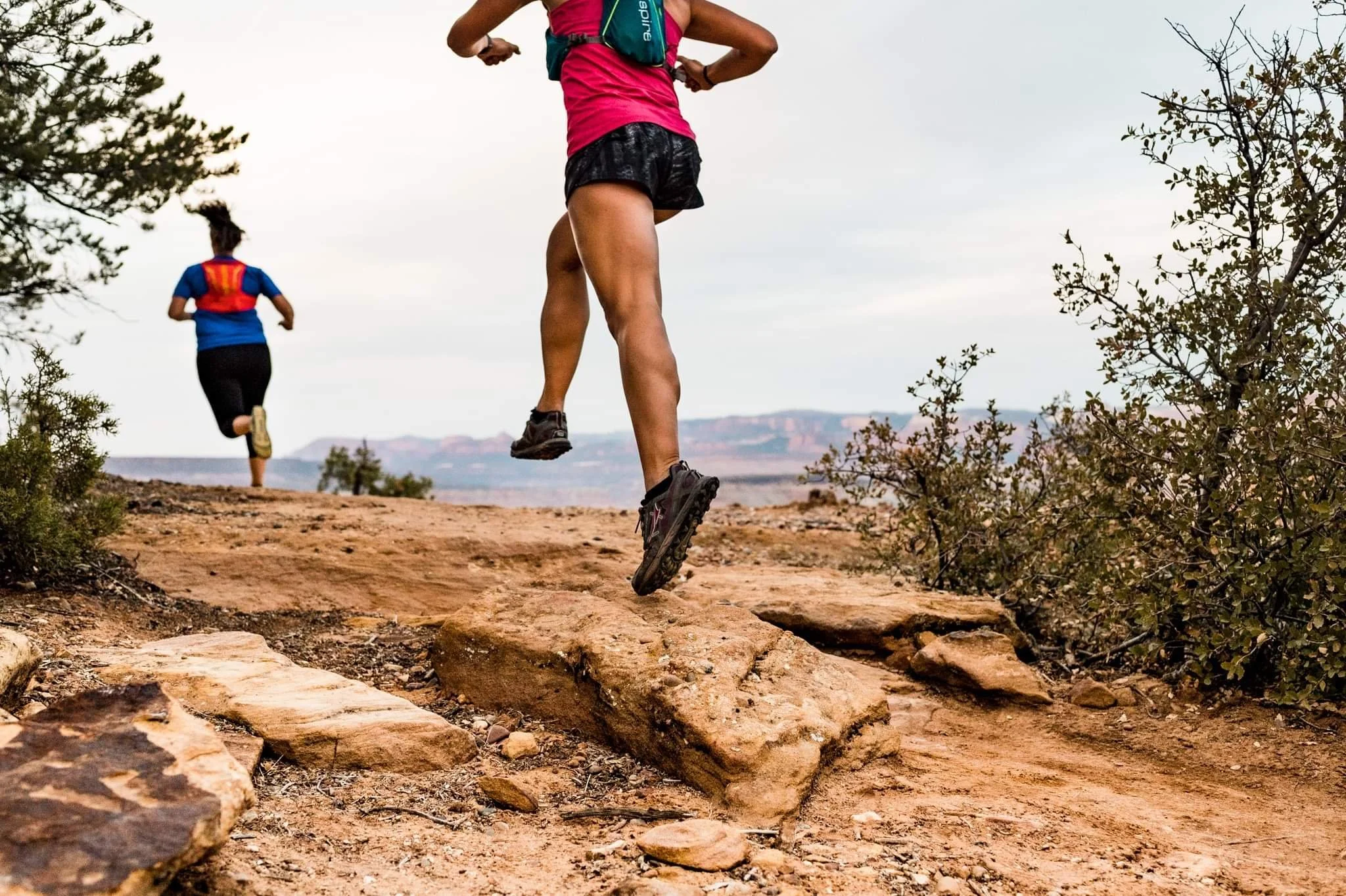 Two people trail running on a rocky path in a desert landscape.