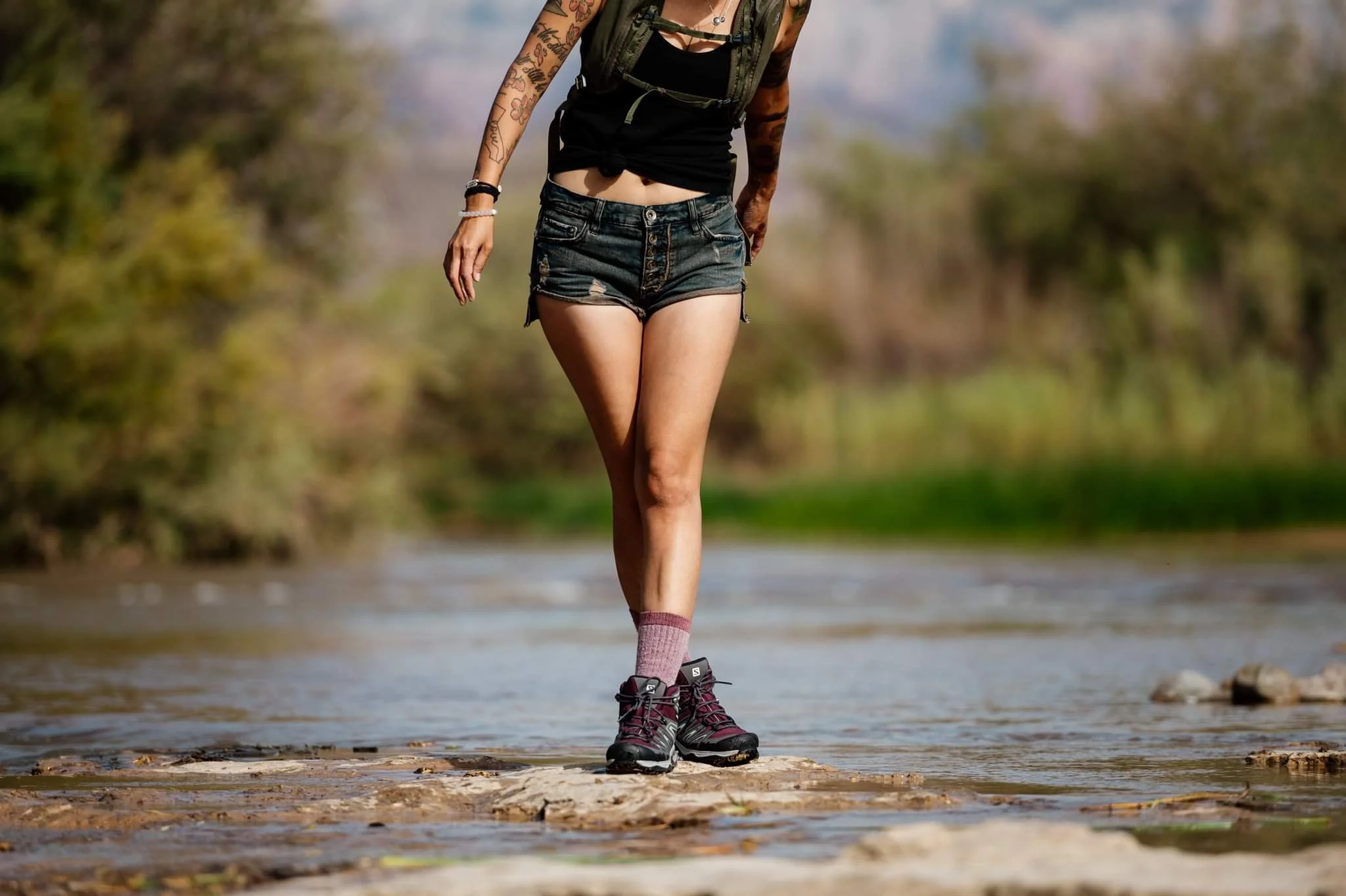 A person stands on rocks in a shallow river, wearing a black tank top, denim shorts, purple socks, and hiking boots.