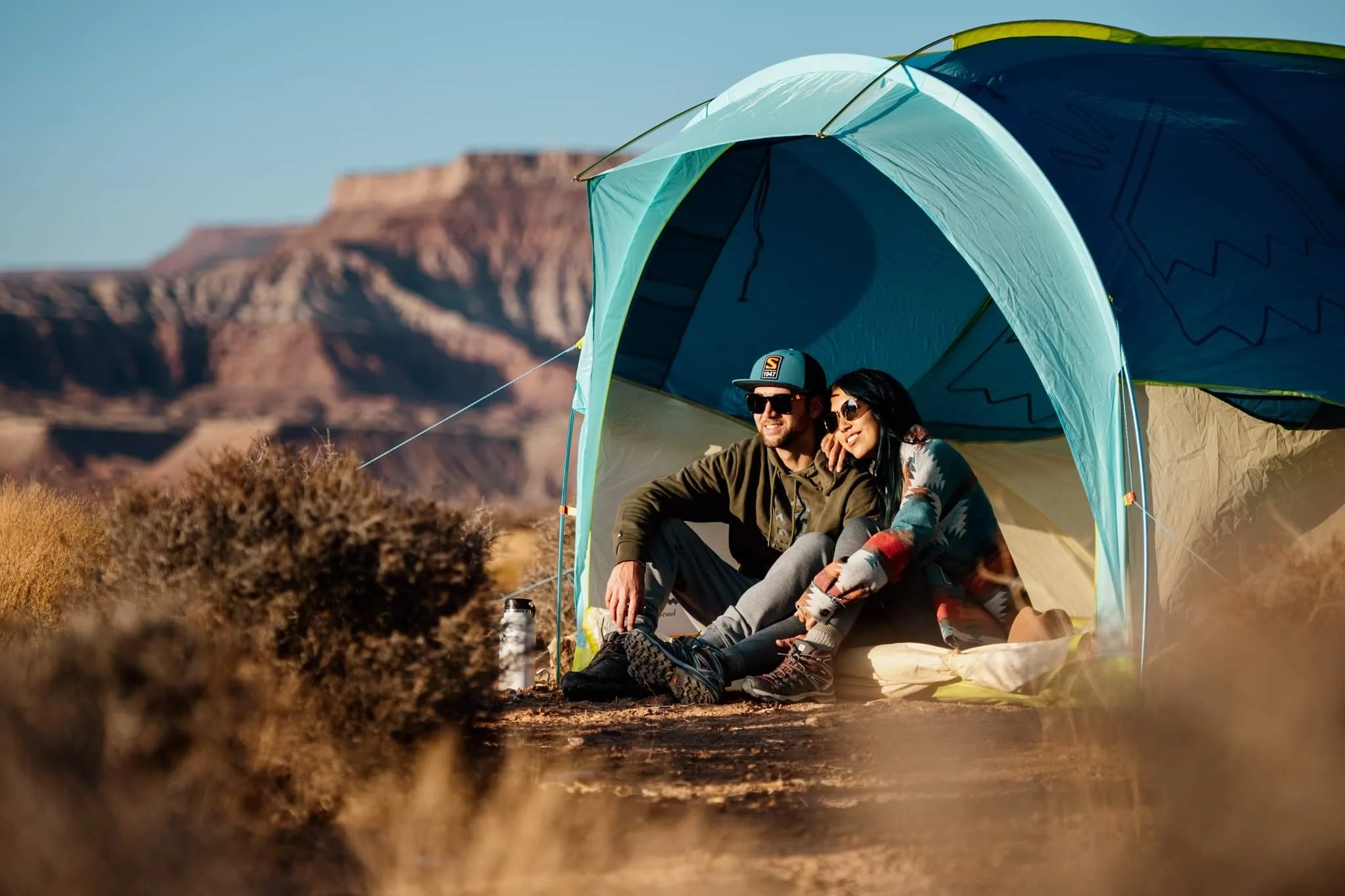 Couple sitting outside a tent in a desert landscape with mountains in the background, enjoying the outdoors.