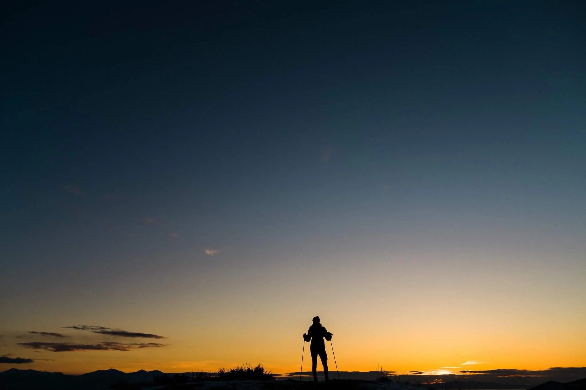 Silhouette of a person hiking with poles against a sunset and mountain landscape.