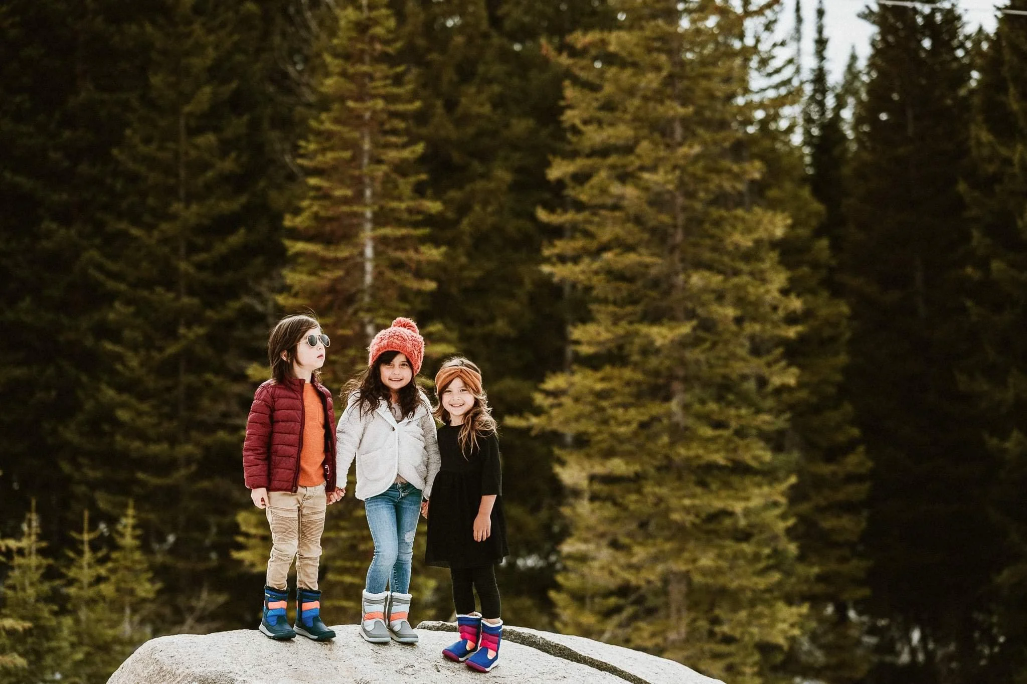 Three children standing on a rock, surrounded by evergreen trees, wearing winter clothing and boots.